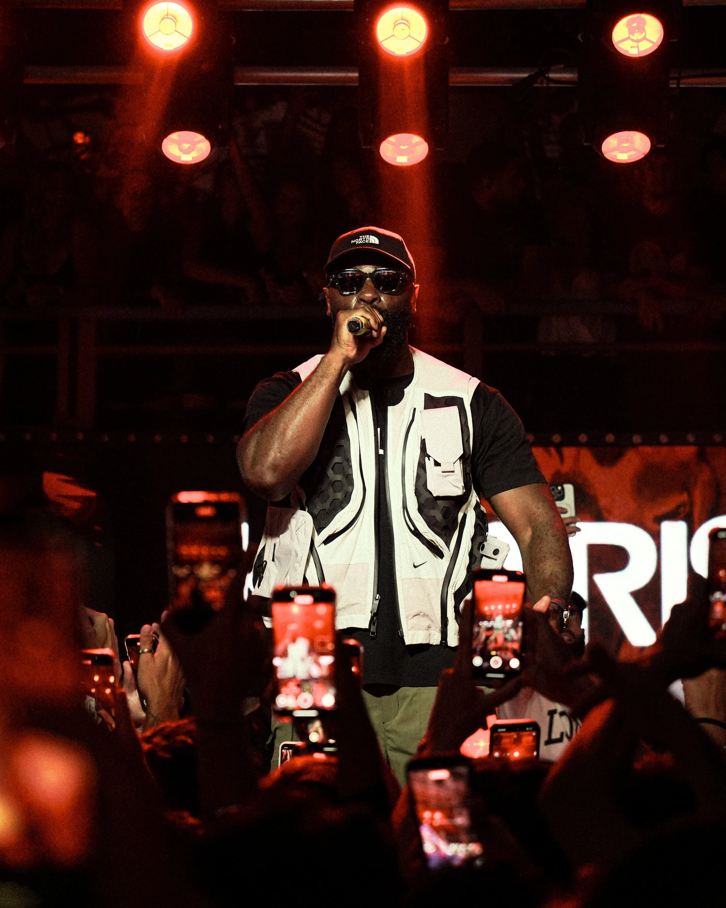 A man wearing sunglasses, a black cap, and a black and white shirt is on stage holding a microphone. Stage lights shine down from above, and there is a crowd in front holding up phones to record the moment.
