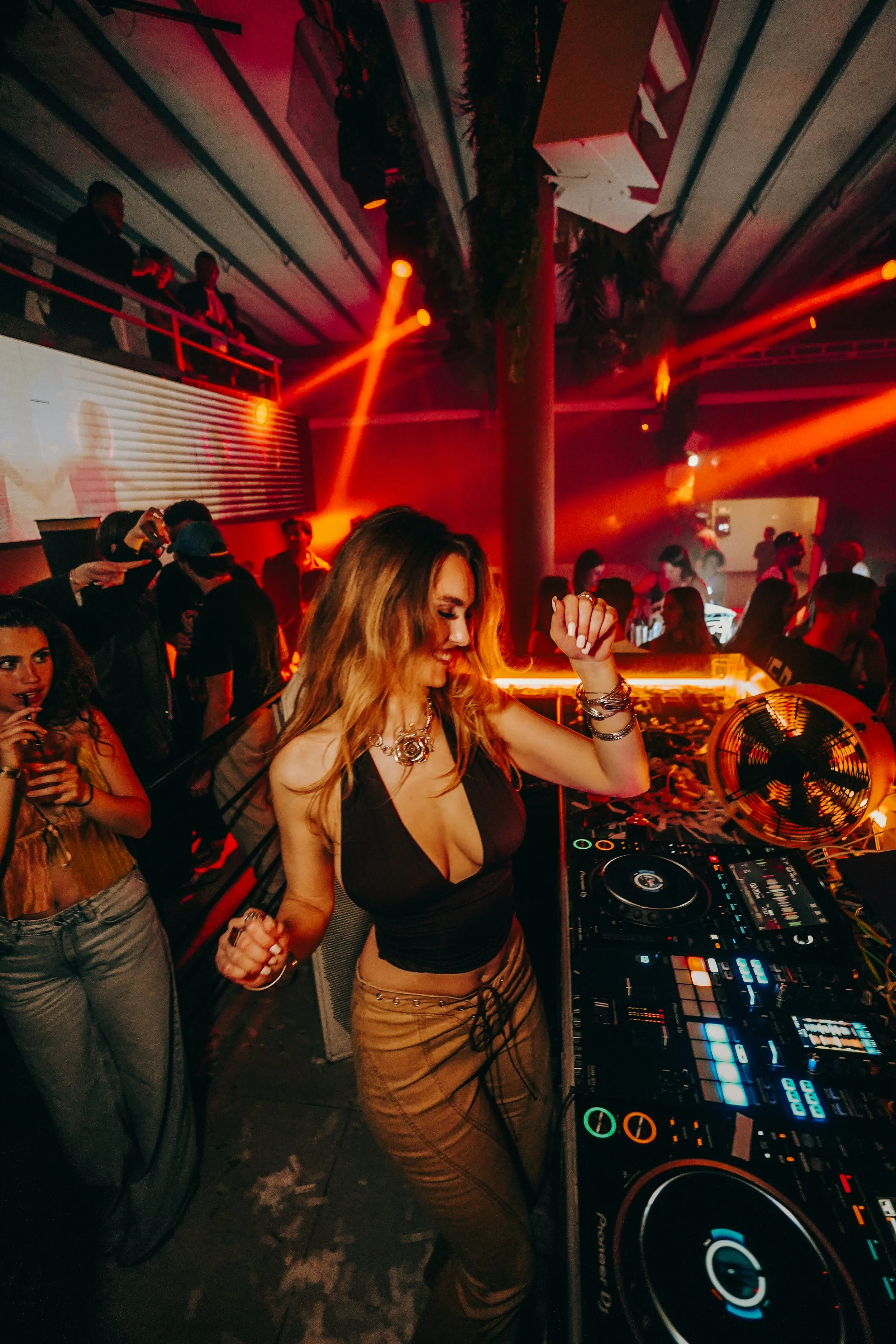 A DJ woman with long blonde hair dancing and mixing music at a nightclub with people in the background, red and orange lighting, and a DJ controller in front of her.