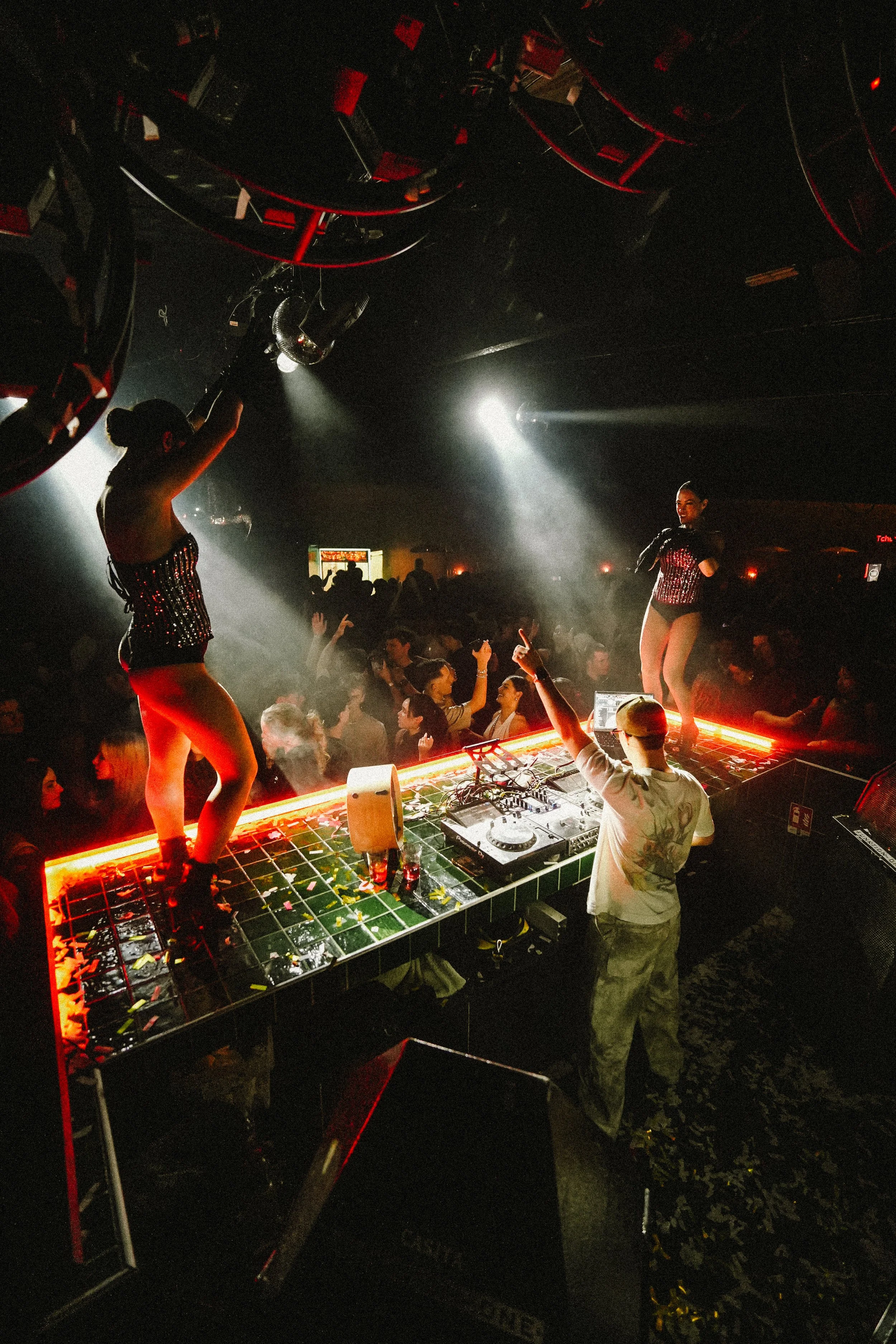 A DJ performing on a decorated platform at a nightclub with two female dancers in sparkling outfits. The crowd is dancing under low lighting with spotlights.