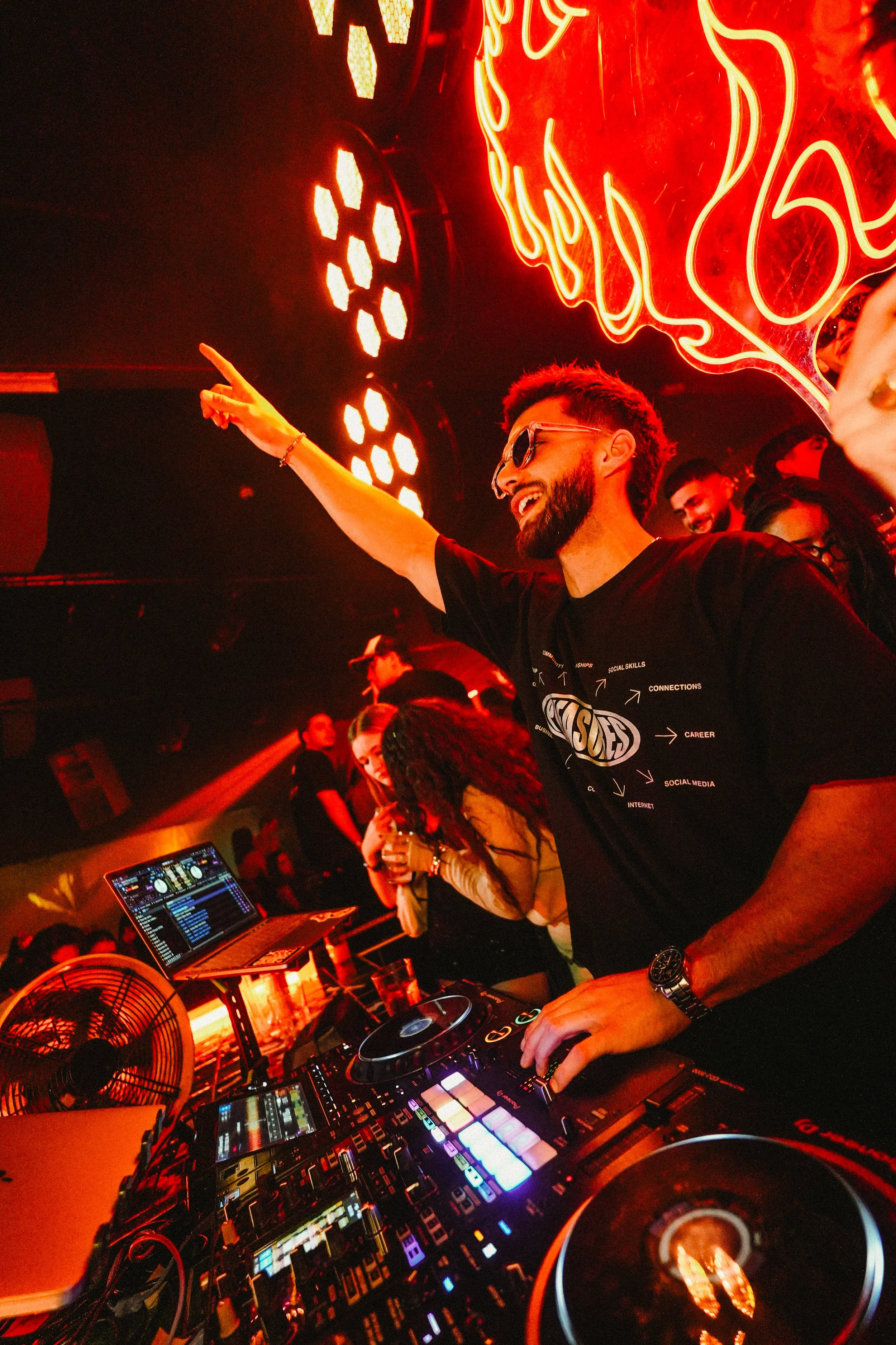A man with sunglasses and a beard, smiling and dancing, at a nightclub with bright red neon lights on the wall behind him. He is DJing with a control panel in front of him, surrounded by other club-goers enjoying the party.