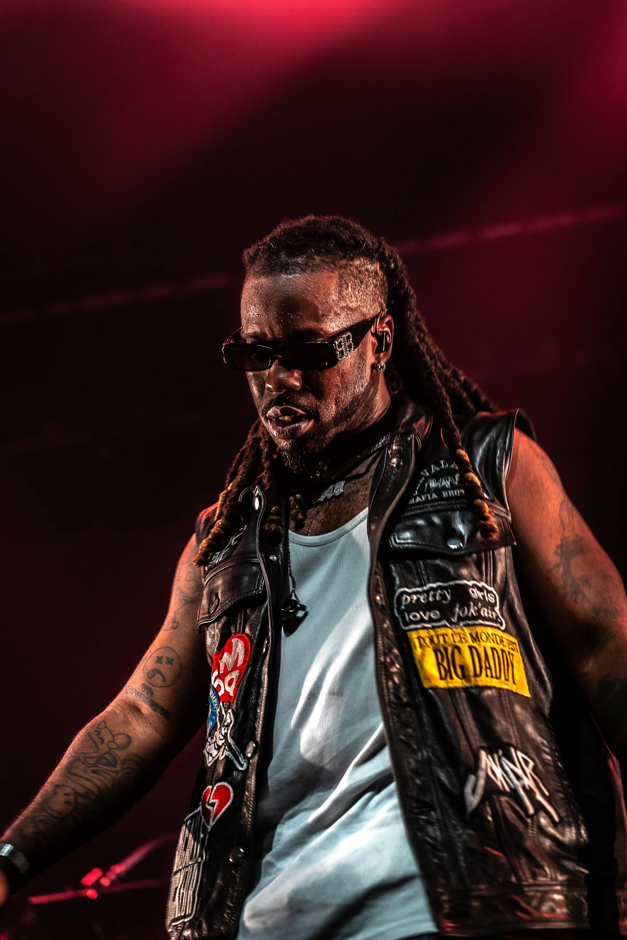 A man with long dreadlocks, wearing sunglasses, tattoos, a white t-shirt, and a black leather vest decorated with patches, performing on stage with red lighting.