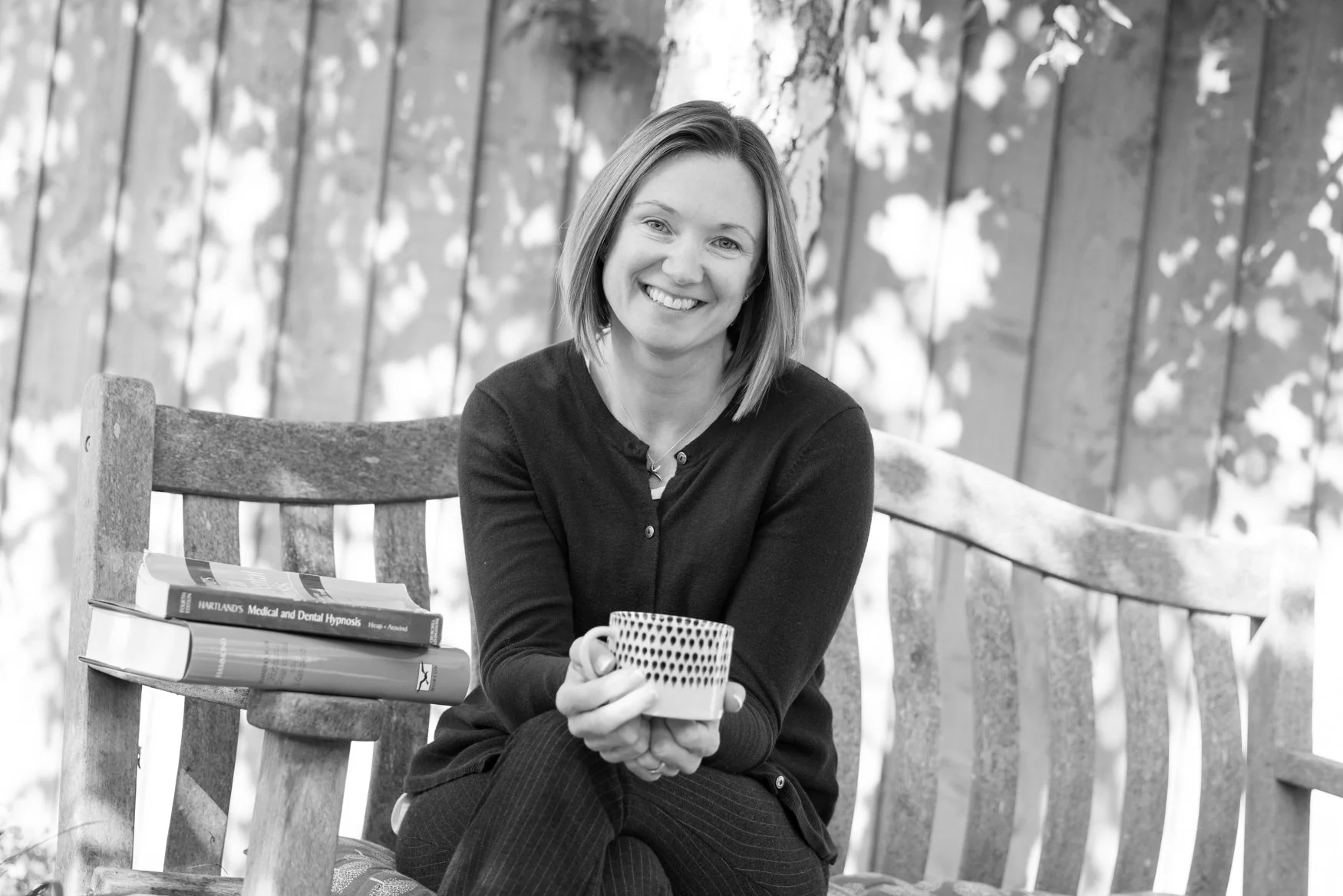 A black-and-white photo of a woman sitting on a wooden bench outdoors, smiling, holding a cup, with a stack of books nearby and a background of a fenced yard with trees.