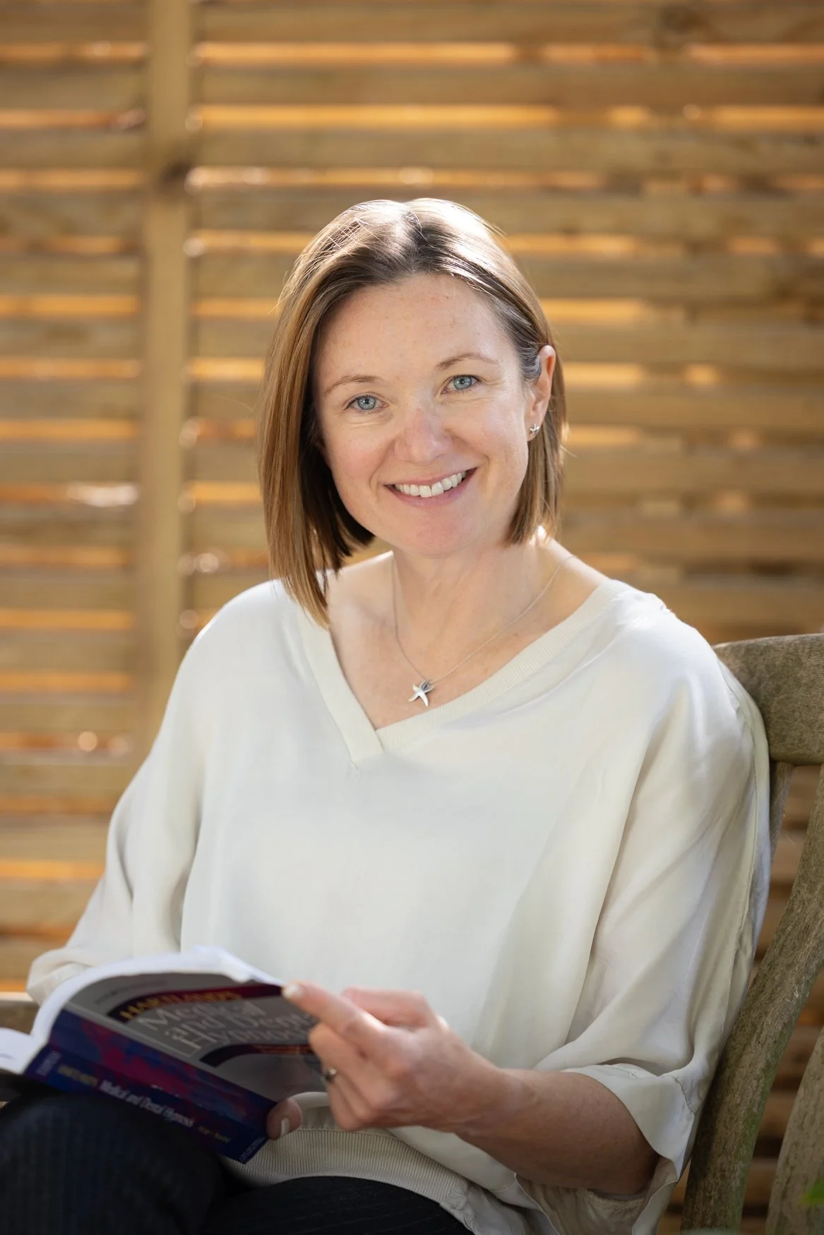 A woman with light brown hair, blue eyes, and freckles smiling at the camera. She is seated indoors, wearing a cream-colored top, a starfish necklace, and earrings. She is holding a magazine or book and sitting in front of a wooden slat backdrop.