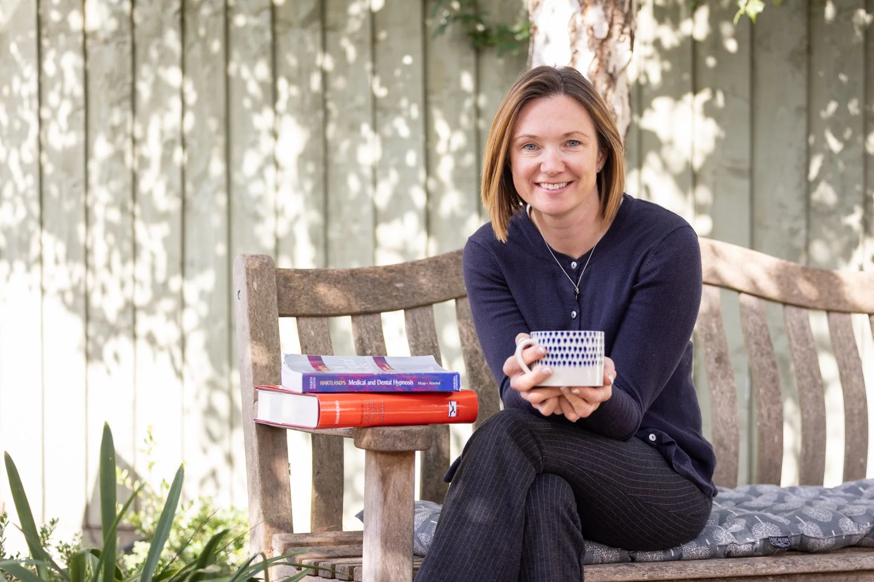 A woman with shoulder-length brown hair, wearing a navy blue cardigan and black pinstripe pants, is sitting outdoors on a wooden bench with a gray cushioned seat. She is smiling and holding a white mug with a blue pattern. Beside her on the bench are three books and a red notebook. The background has a light-colored wooden fence and a tree, with sunlight filtering through the leaves.