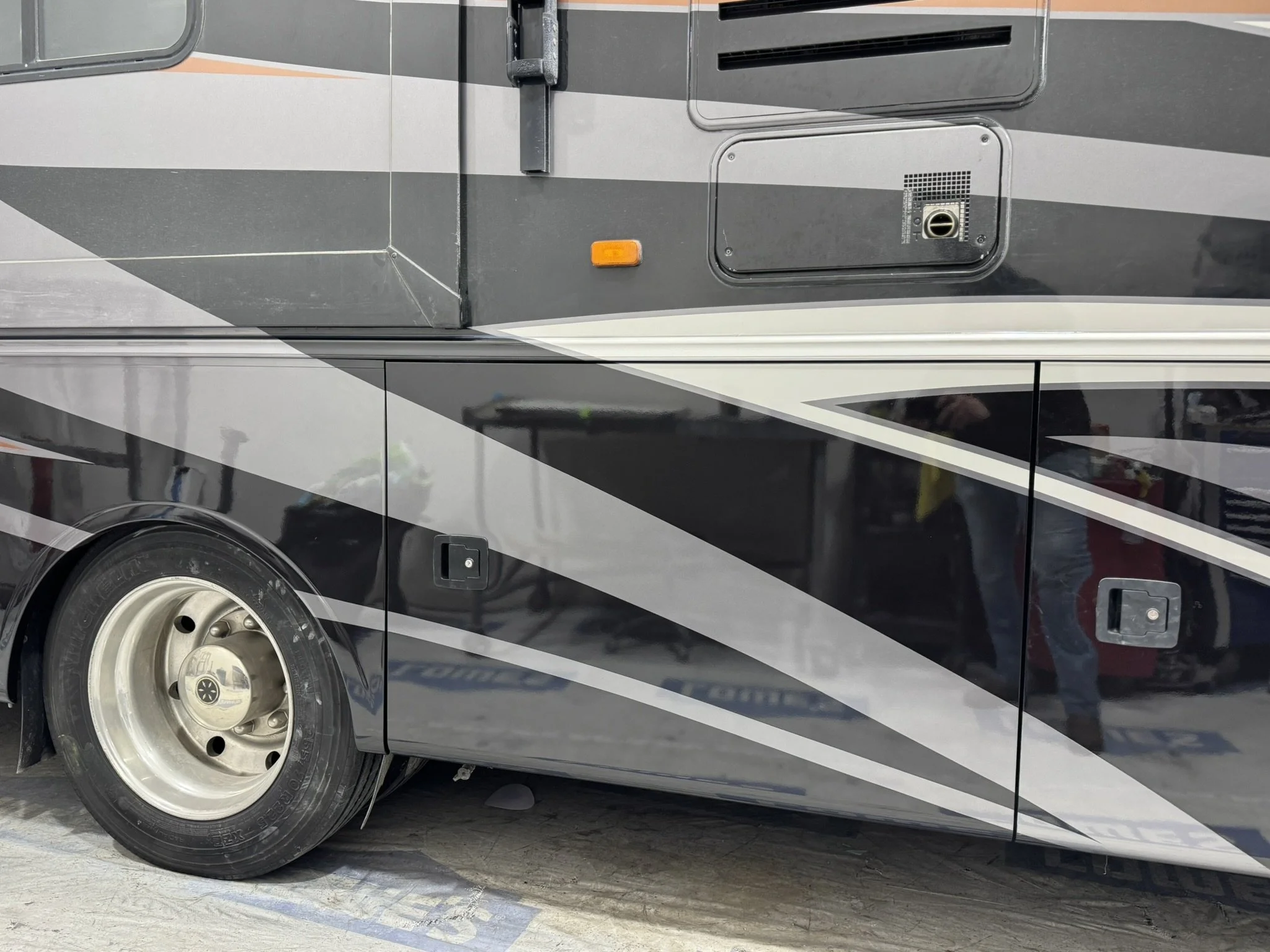 Close-up of a section of a black and gray RV or bus with a wheel, small orange marker light, vent, and access panels on the side, parked on a concrete surface.