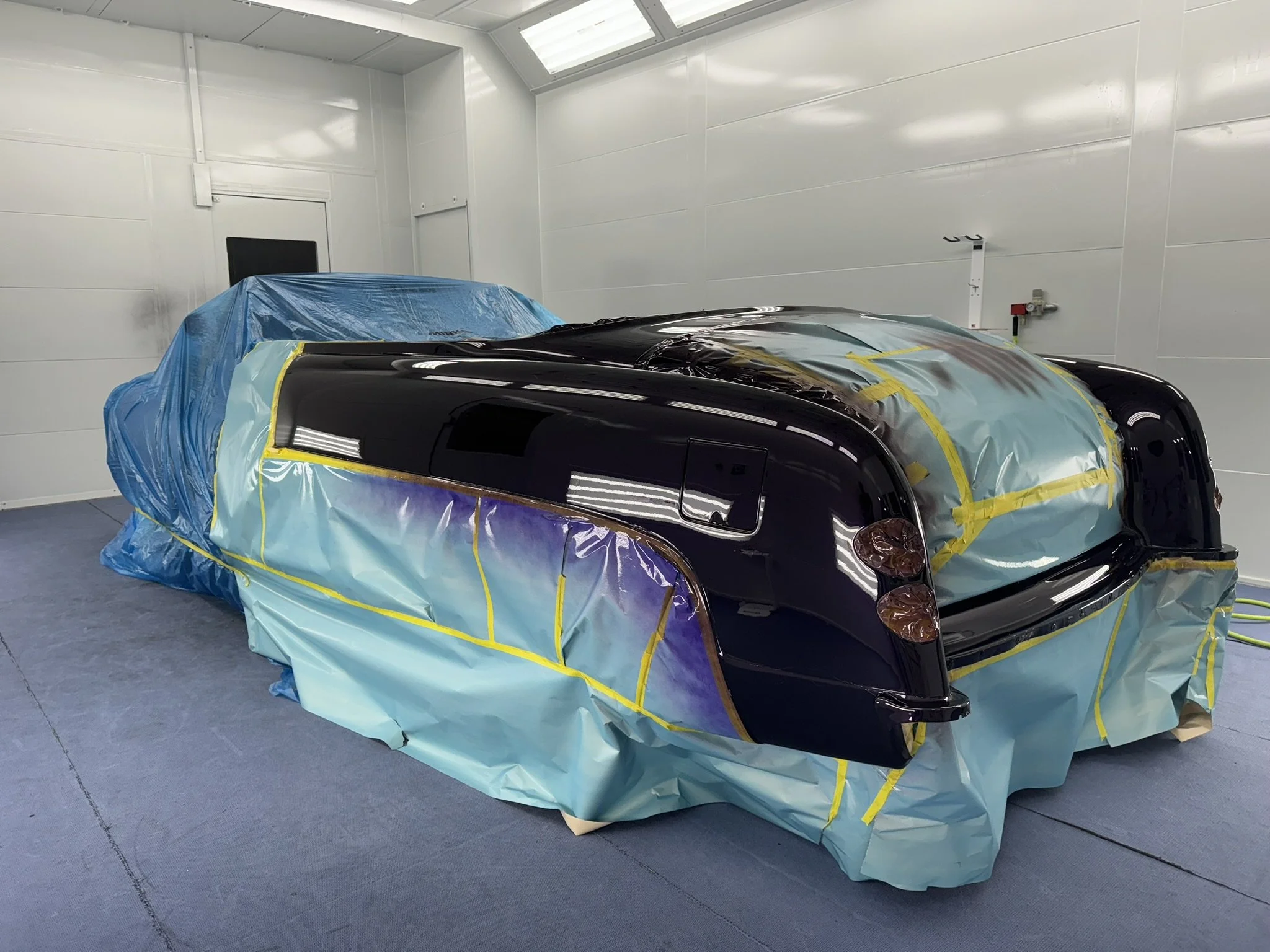 A black sports car in a paint booth, covered with protective blue and yellow plastic sheets, preparing for painting.