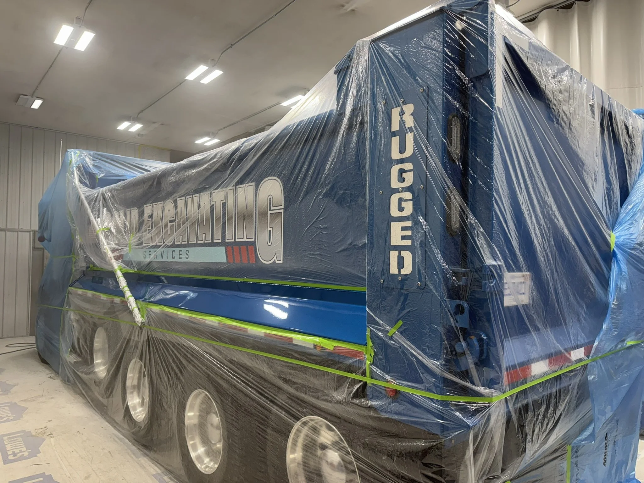 A large truck covered in blue paint and graphic decals, wrapped in clear plastic for protection, inside a warehouse or factory building.