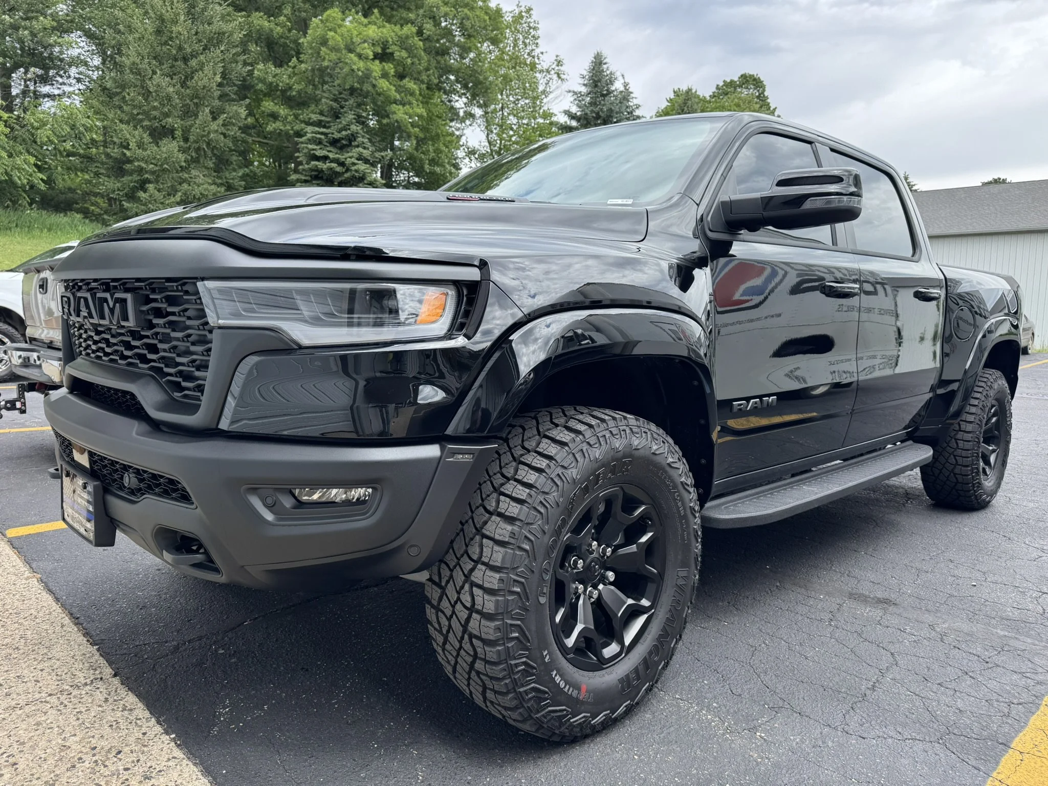 Front and side view of a black RAM pickup truck with large off-road tires parked in a lot with trees and a building in the background.