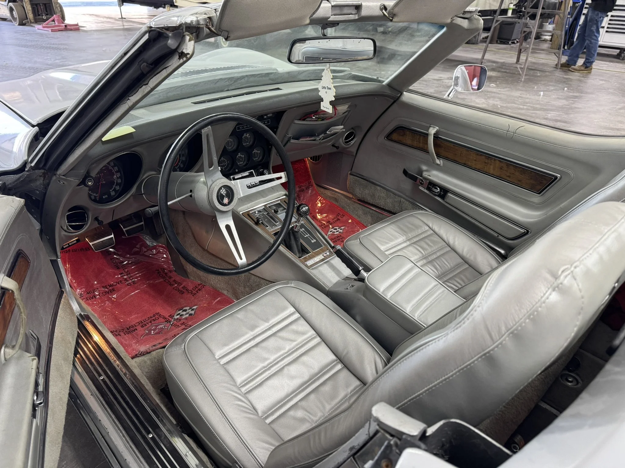 Interior of a vintage convertible car with gray leather seats, dashboard, and steering wheel, parked inside a garage or workshop. The dashboard has various gauges, and the windshield has a hanging air freshener shaped like a tree.