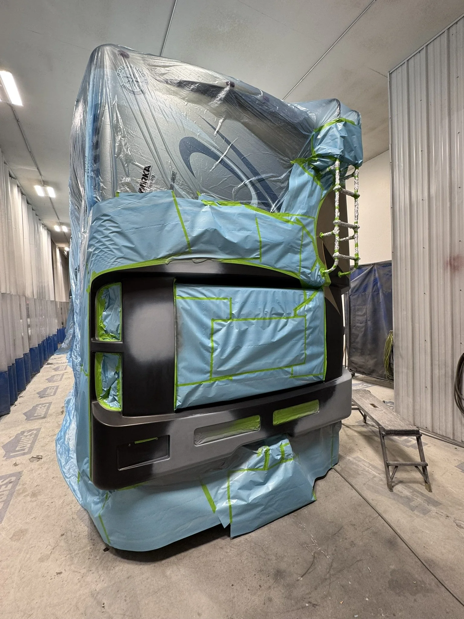 Large machine wrapped in light blue protective covering and plastic sheeting, secured with green tape, in an industrial setting.