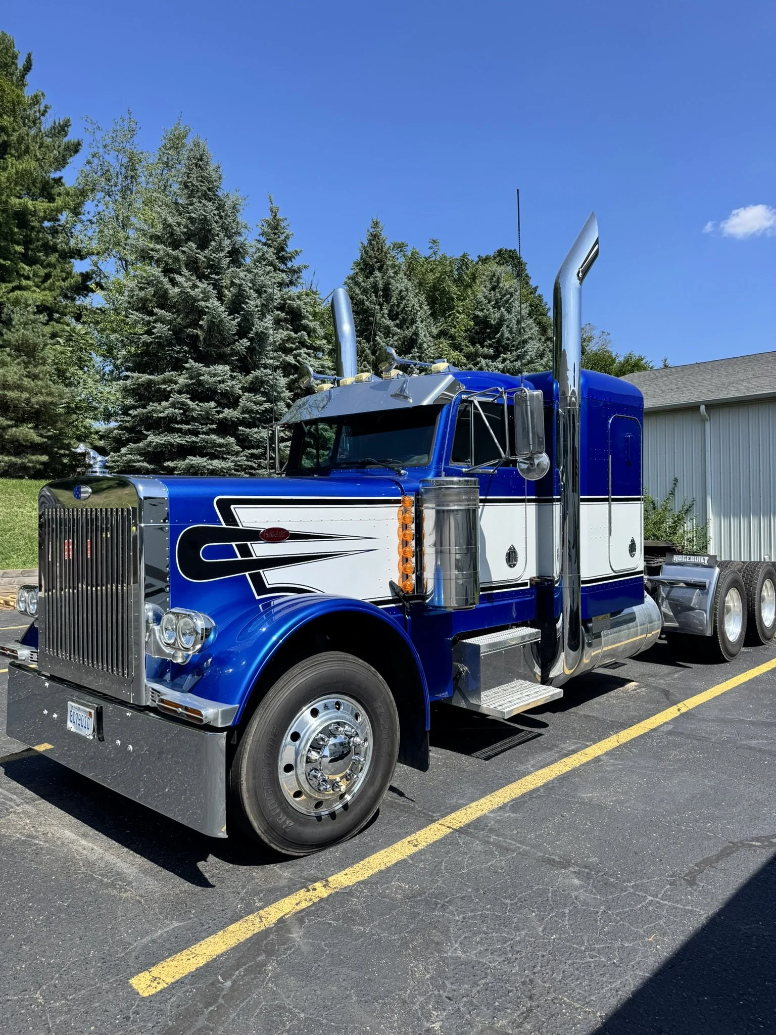 Blue semi-truck parked in a parking lot under a blue sky with trees in the background.