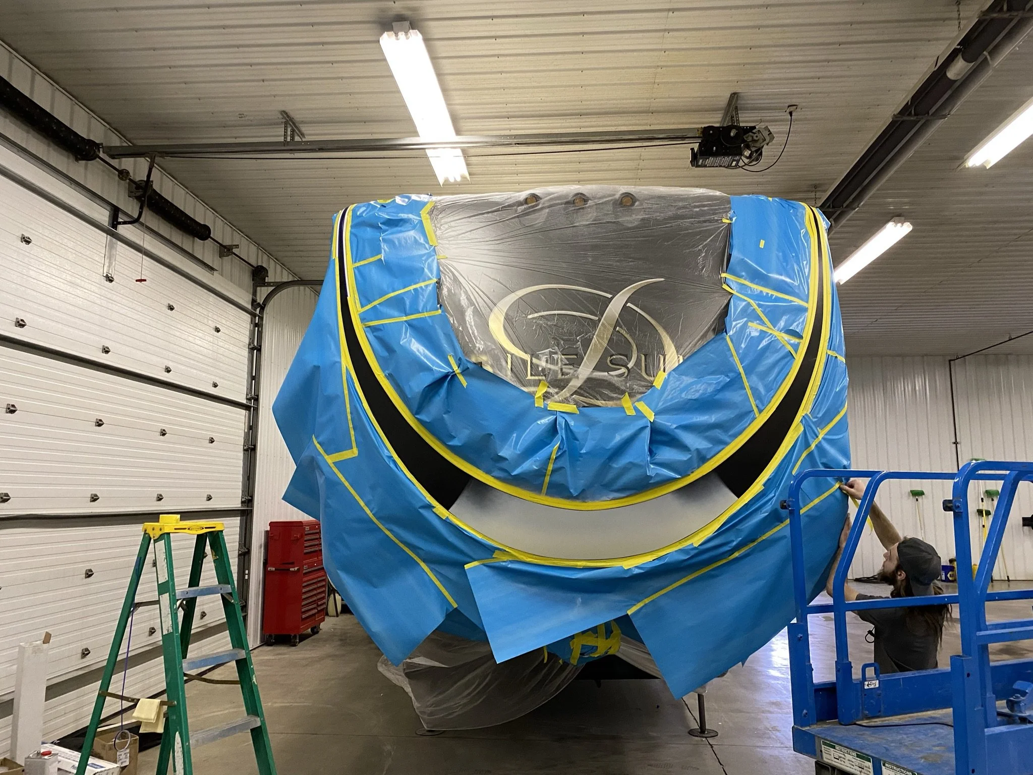 A large object wrapped in a blue and black cover and plastic sheeting, suspended in a warehouse with work ladders, red toolboxes, and a person inspecting the setup.