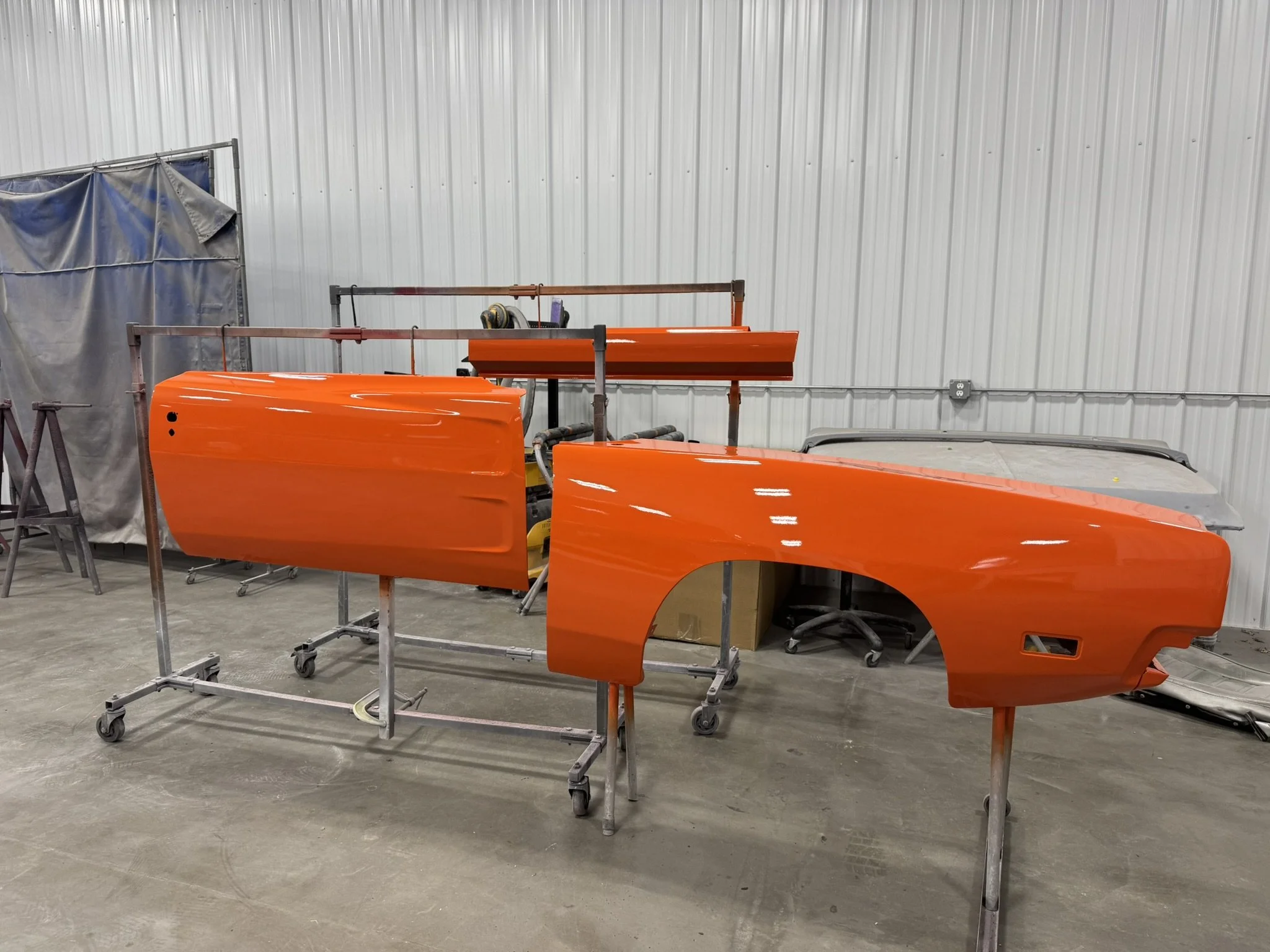 Orange car parts and panels are being painted or assembled inside an industrial workshop with a corrugated metal wall in the background.