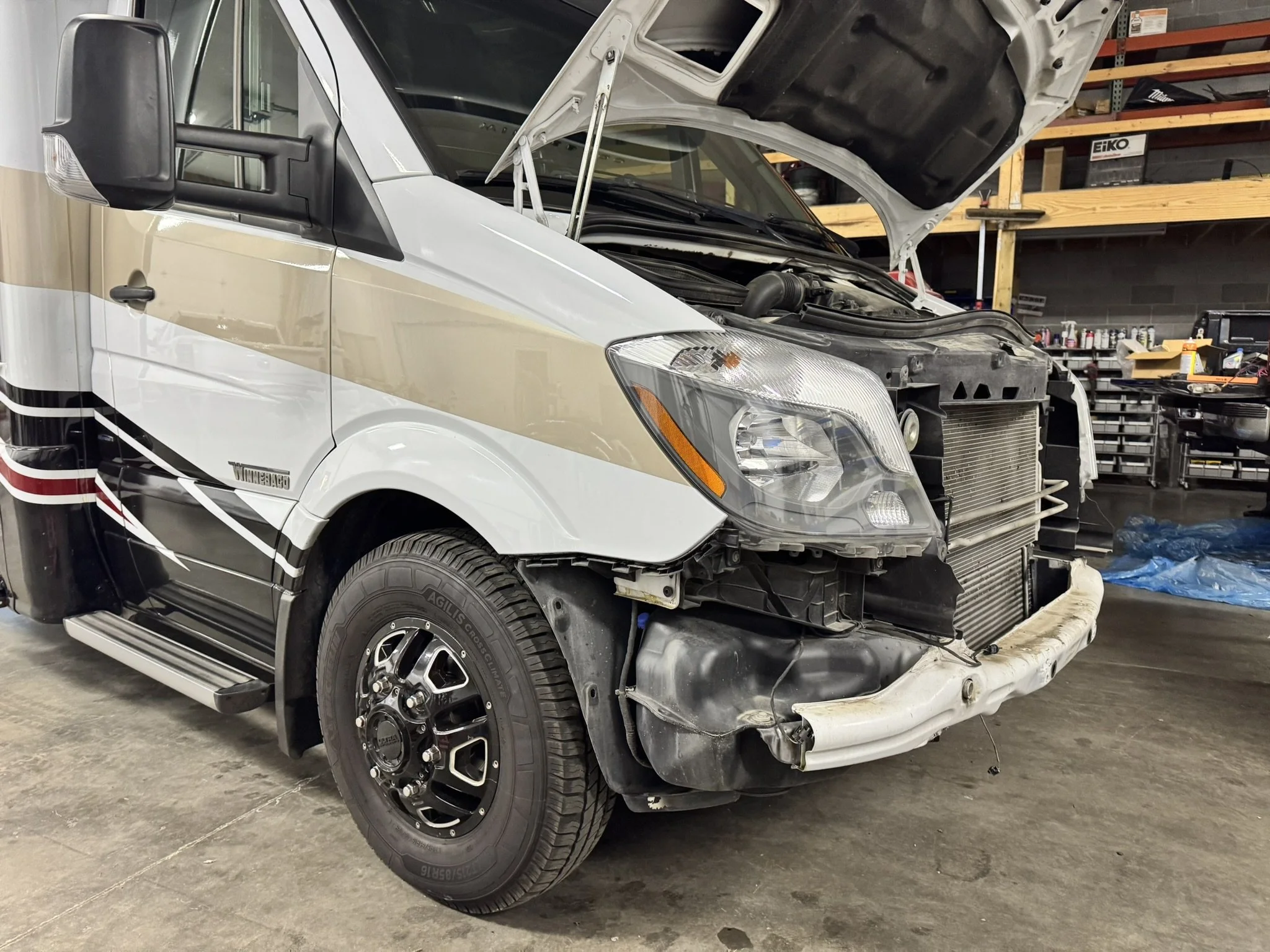 A white and beige RV with its front bumper and grille removed, exposing the radiator and engine components, parked inside a workshop with shelves and tools in the background.