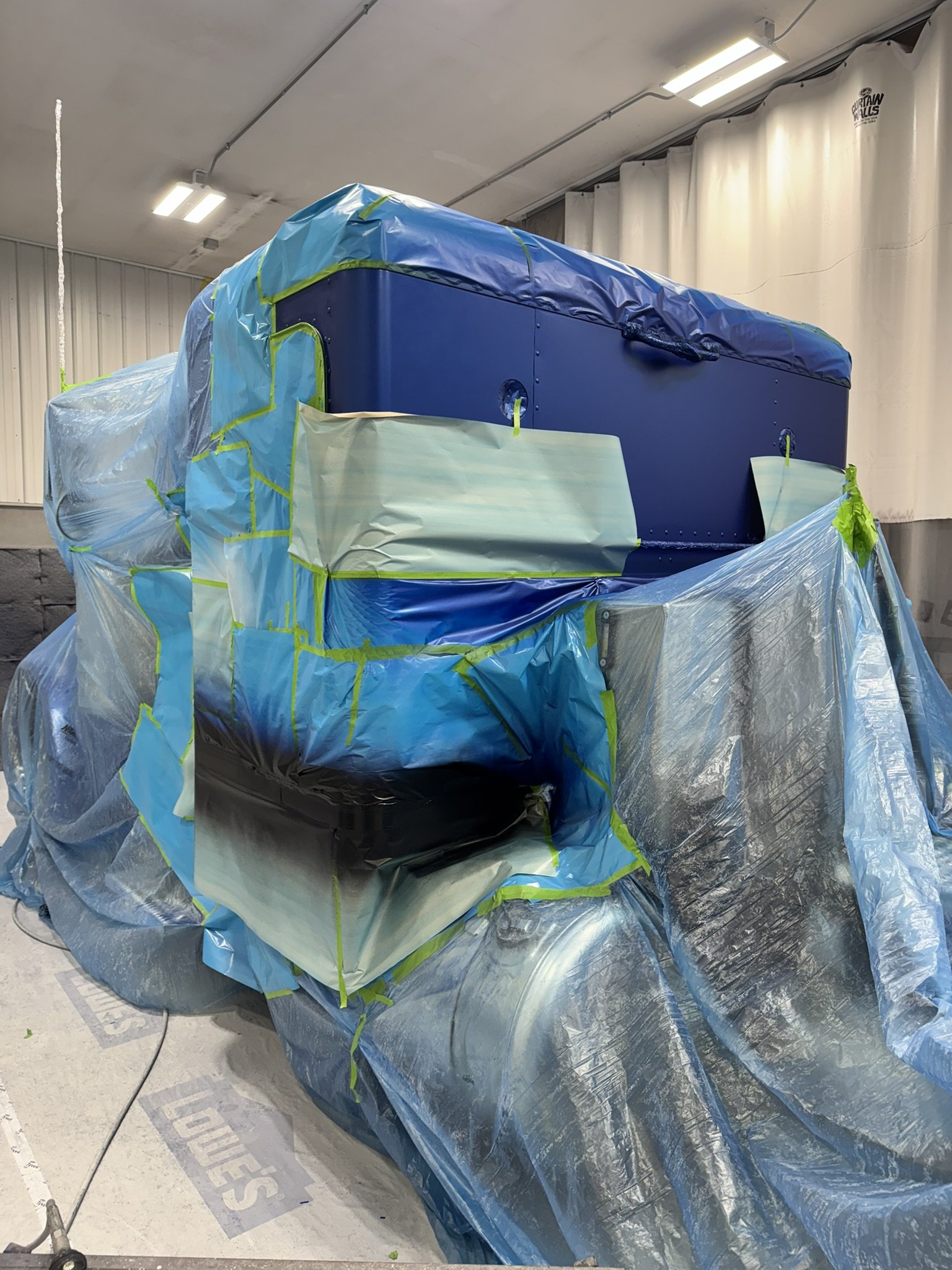 A vehicle covered in blue and silver protective wrapping, with some green tape, inside an indoor space with white curtains and ceiling lights.