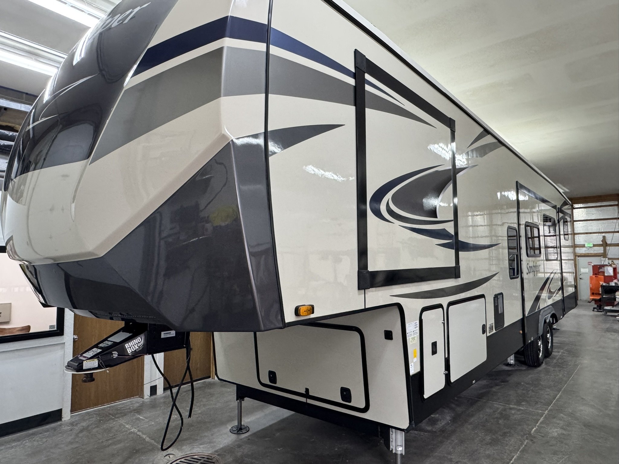 Side view of a large white and gray RV with black and blue graphics, parked indoors with a concrete floor and ceiling.