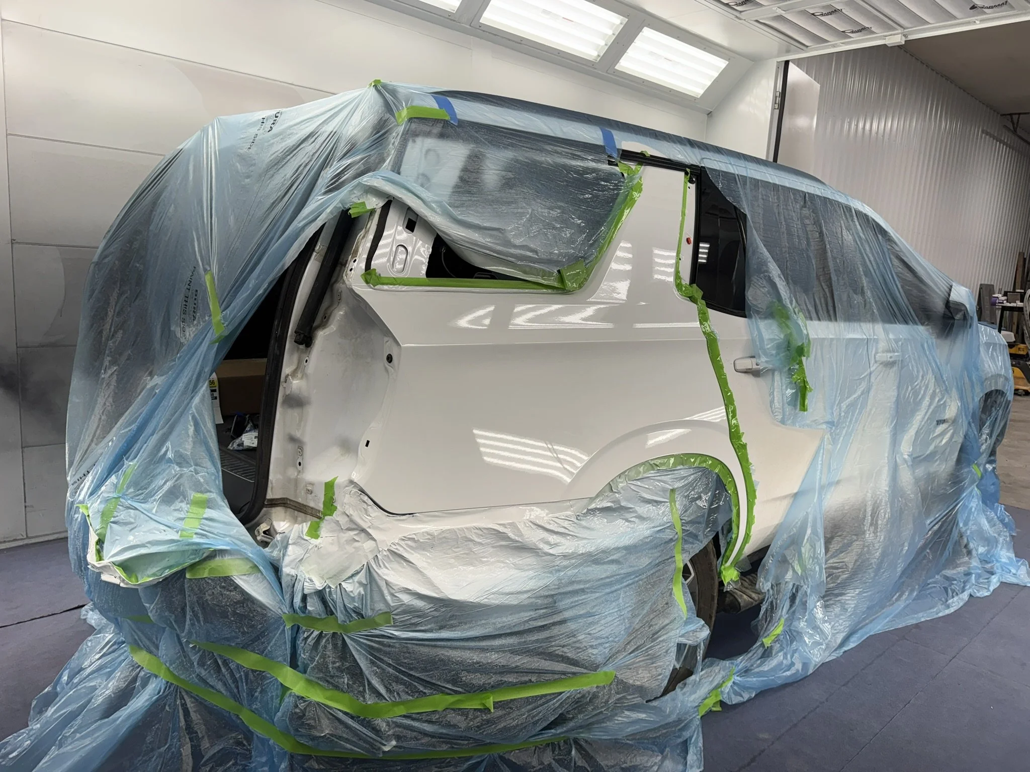 A white vehicle is covered with blue plastic and green painter's tape, indicating it is being prepared for painting or repair in a workshop with white walls and ceiling lights.