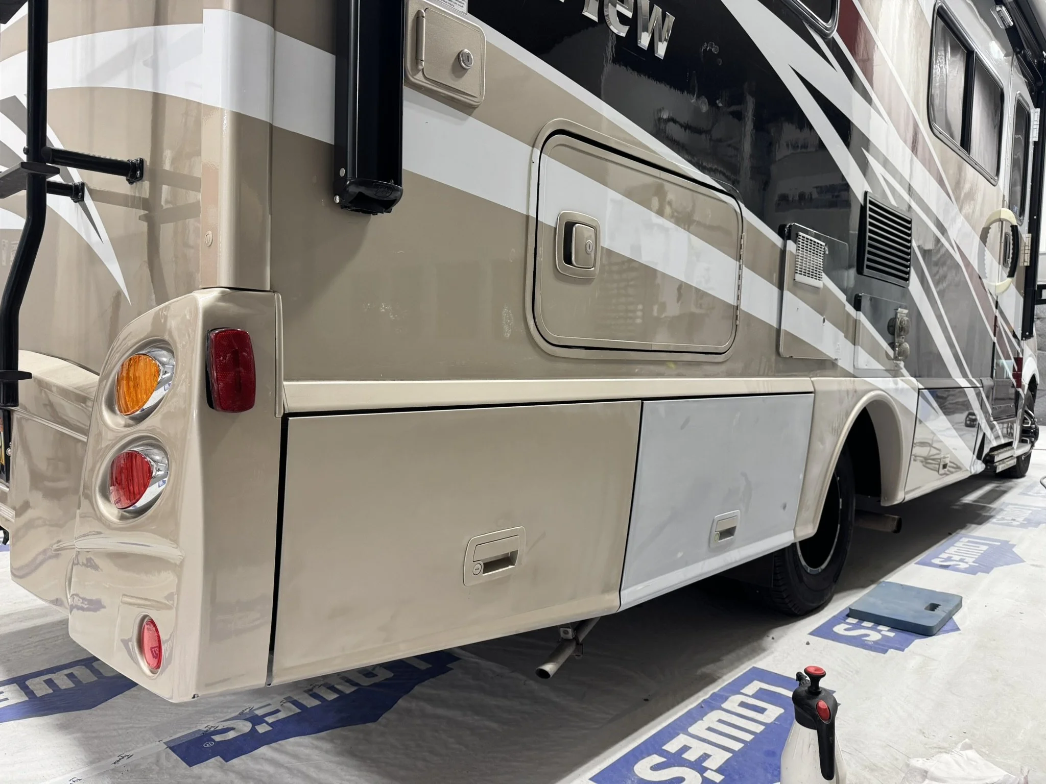 A beige and black recreational vehicle (RV) parked indoors, with a door, windows, vents, and tail lights visible, along with a black mechanism and a red button on the floor.