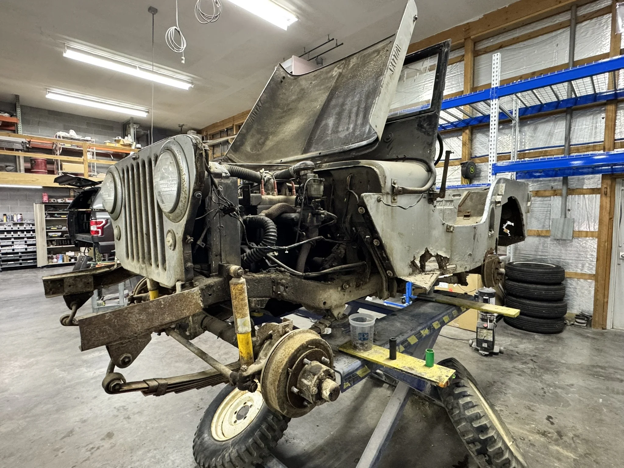 A vintage Jeep vehicle in a workshop, with the front hood open and the front end disassembled, showing the engine and internal parts. The workshop has shelves with tools and supplies, and tires stacked on the side.