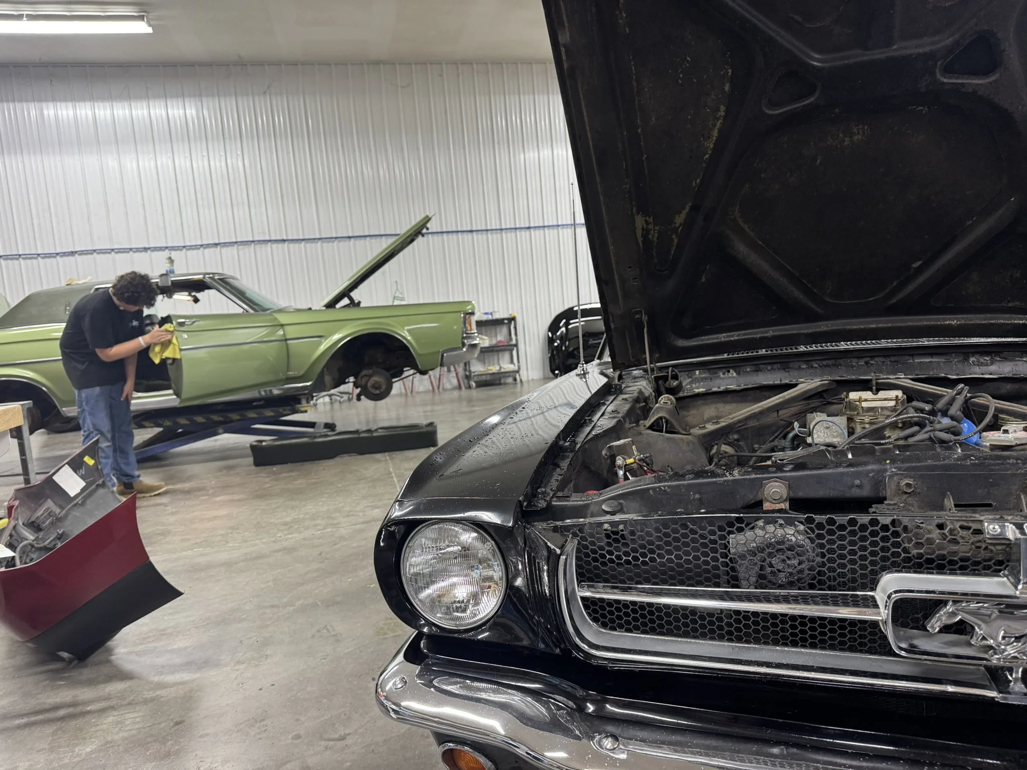 A mechanic is working on a black car with the hood open inside a garage, with a green classic car elevated on a lift in the background.