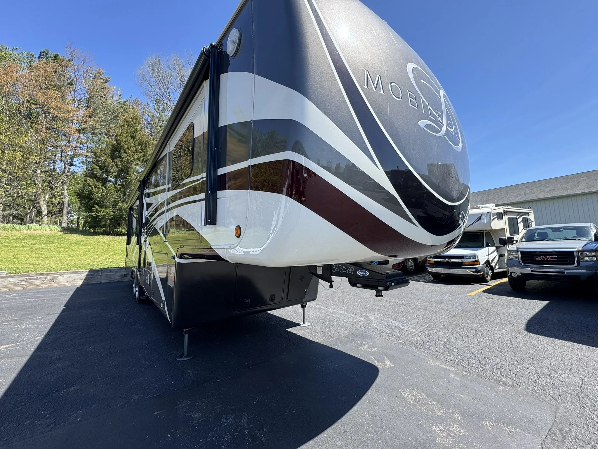 Front view of a black, white, and gray mobile home or RV parked on a paved lot, with trees and other vehicles in the background.
