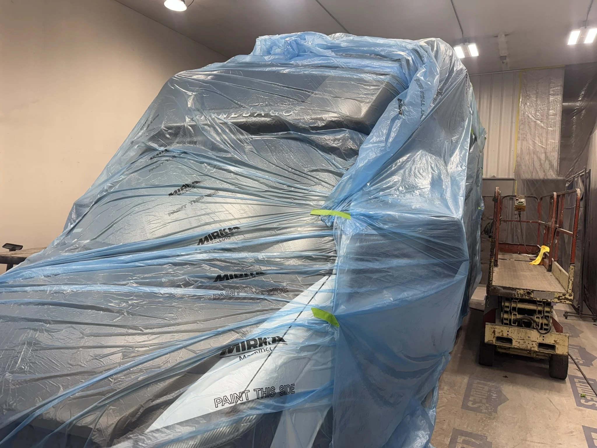 A large object covered with a blue plastic tarp labeled 'Paint This Side' inside a room with unfinished walls and ceiling. There is a red scissor lift and workshop tools nearby.