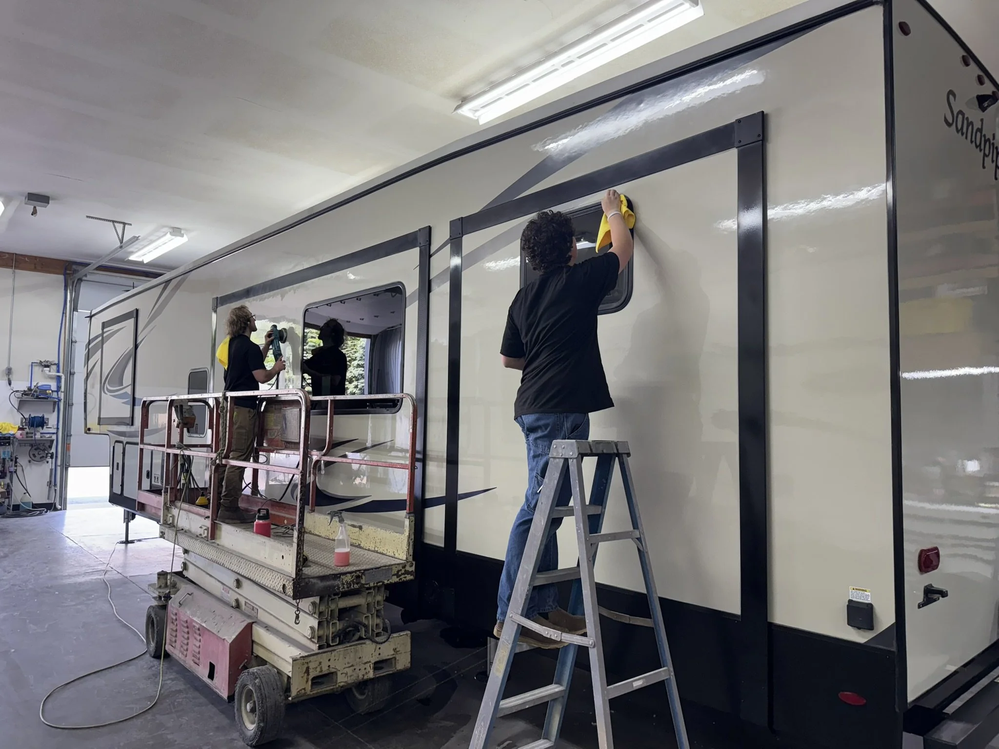 Workers cleaning the side of a large RV in a garage using a yellow cloth and water spray.