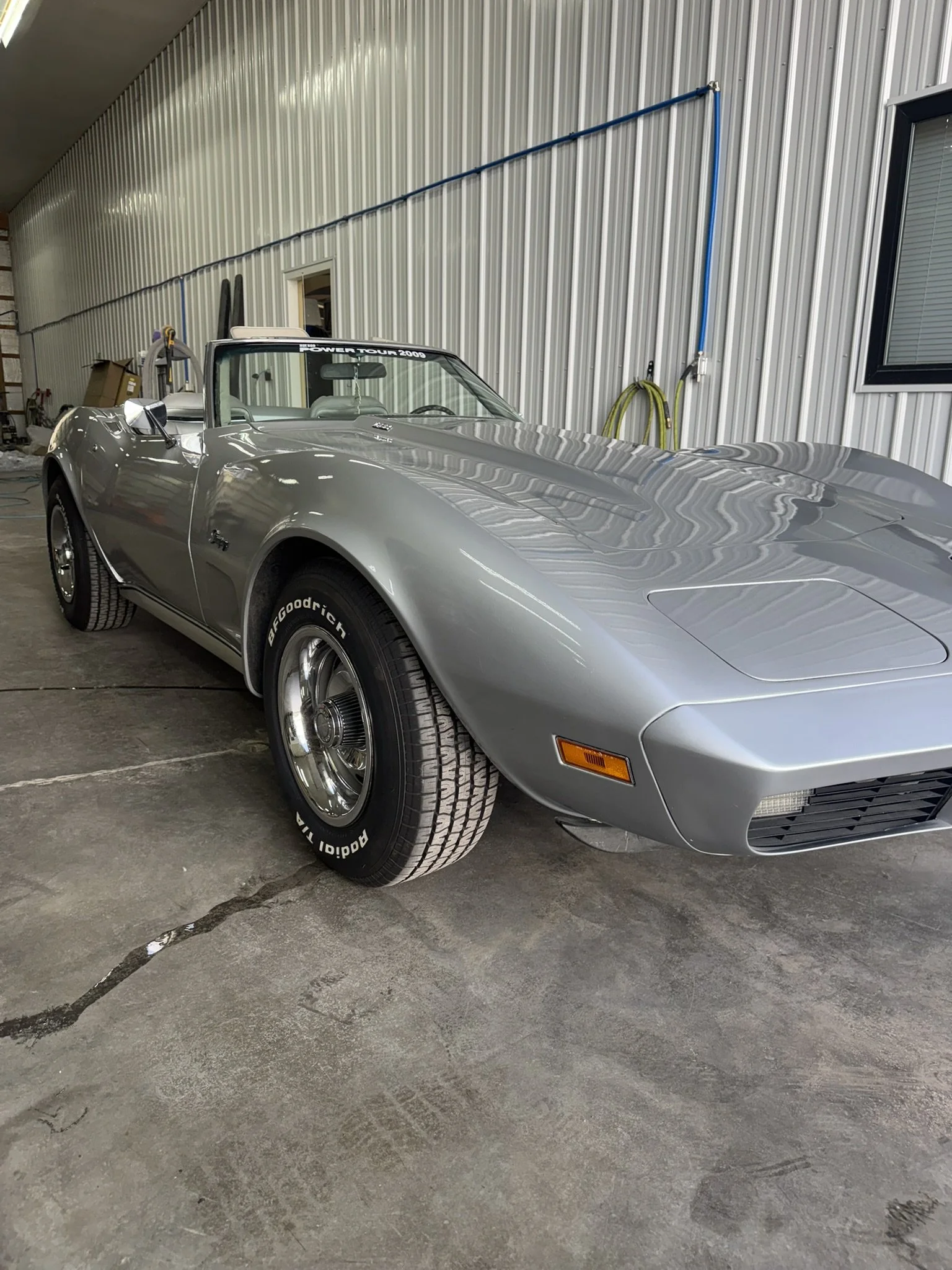 A silver vintage Chevrolet Corvette convertible parked inside a garage with metal walls and concrete floor.