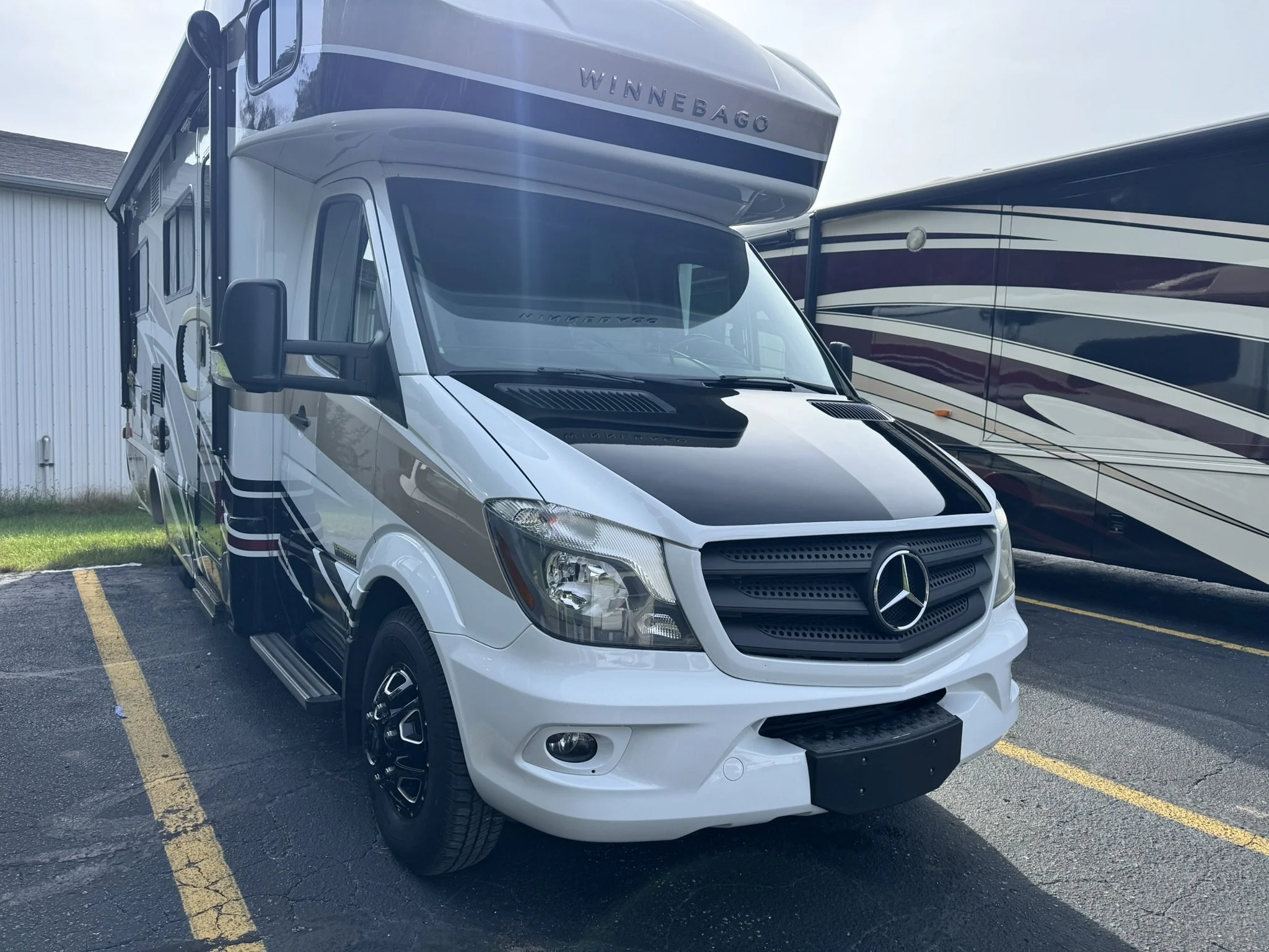 A Winnebago motorhome parked between a large maroon and white RV and a white building with a gray roof, on a paved parking lot with yellow lines.