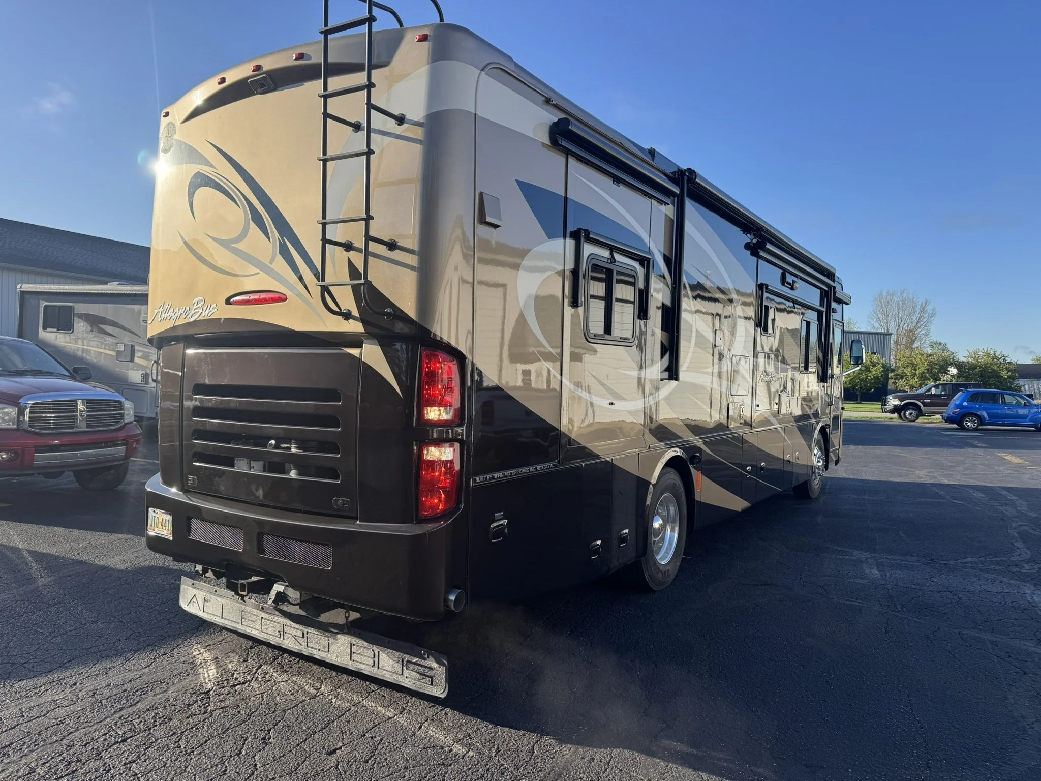 A large motorhome with beige and black exterior parked in a parking lot, with other vehicles and buildings in the background under a clear blue sky.