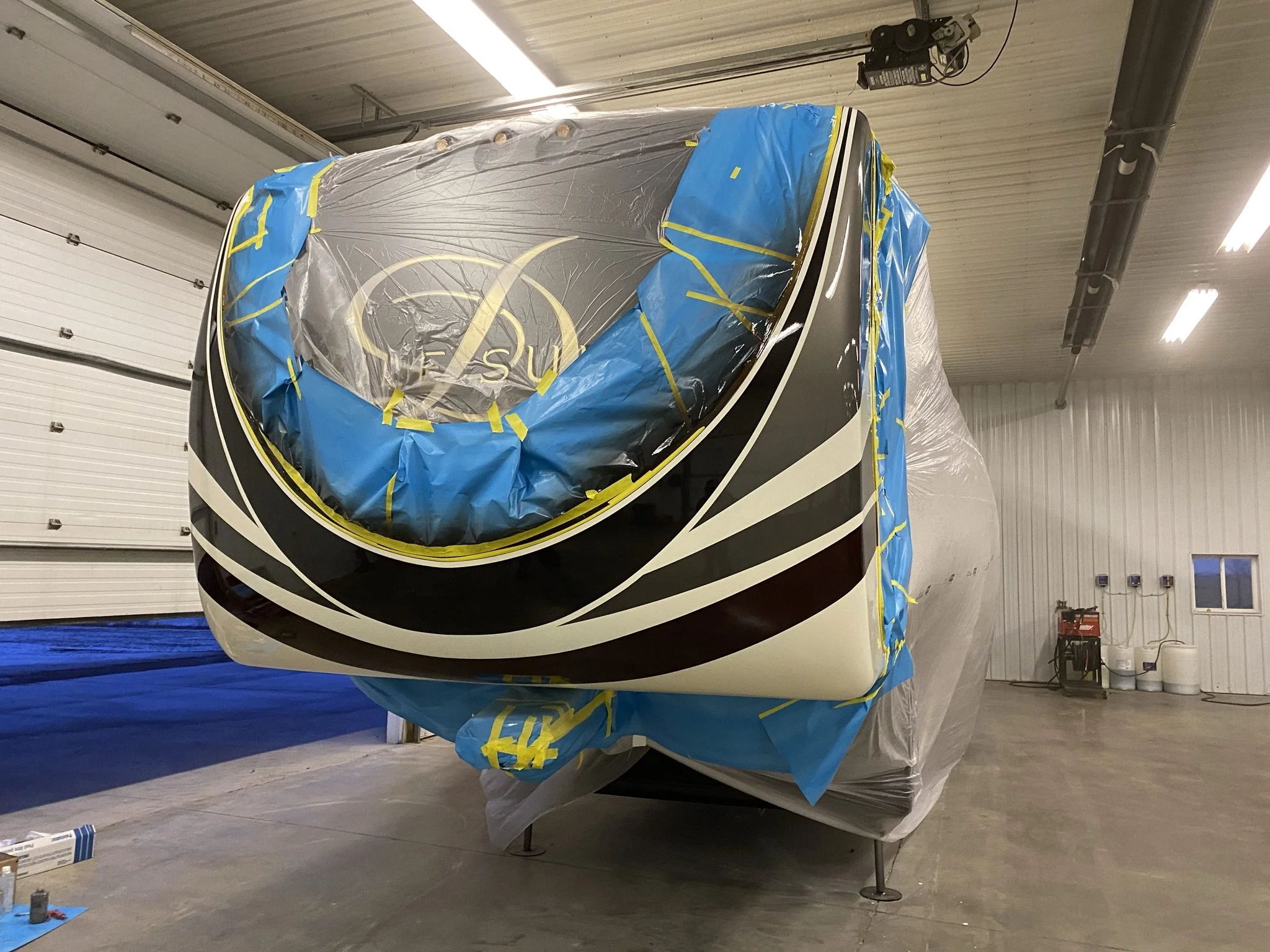 A boat covered with blue and clear plastic sheets with yellow tape, parked inside a large garage or hangar with white walls and ceiling and fluorescent lighting.