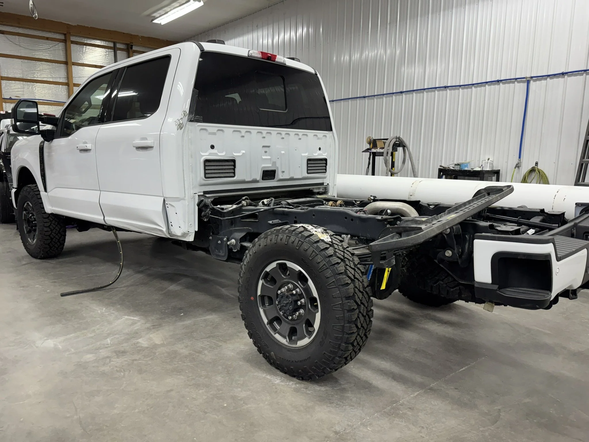 A white pickup truck in an indoor workshop, with the bed and rear panel removed, exposing the chassis and suspension components. The truck is large with off-road tires.