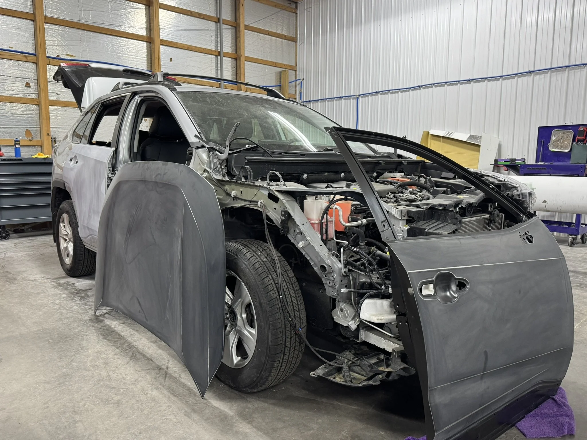 A silver car is in the process of being repaired or assembled inside a workshop or garage. The front of the car is disassembled, exposing the engine and internal components. The car is surrounded by various tools and equipment.