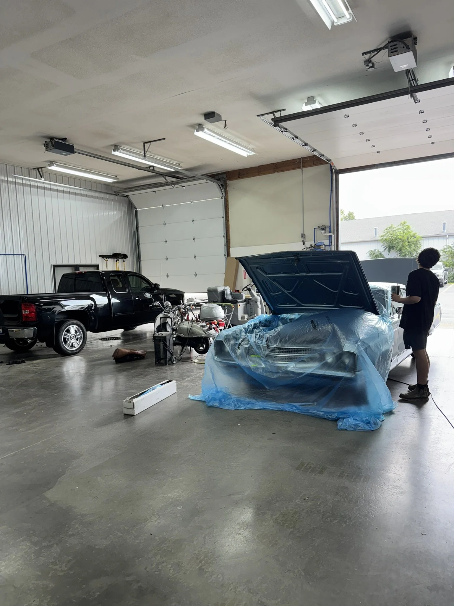 Inside a garage with vehicles and equipment, a woman stands near a silver car with its hood open, covered in blue plastic, while other vehicles and items are present; garage door is partially open letting in outside light.