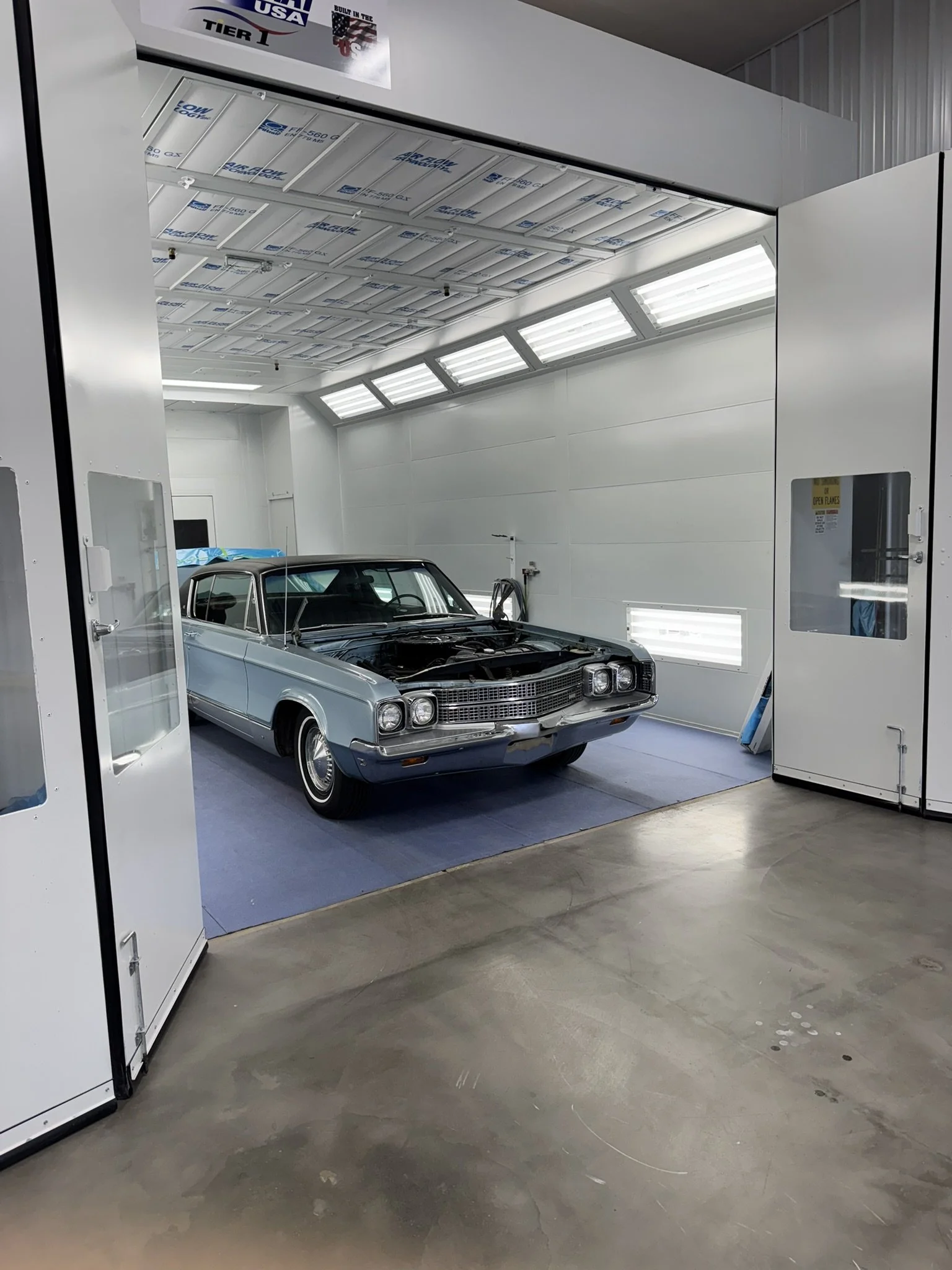 A classic silver car inside a clean, brightly lit automobile paint booth with white walls and ceiling, and a concrete floor.