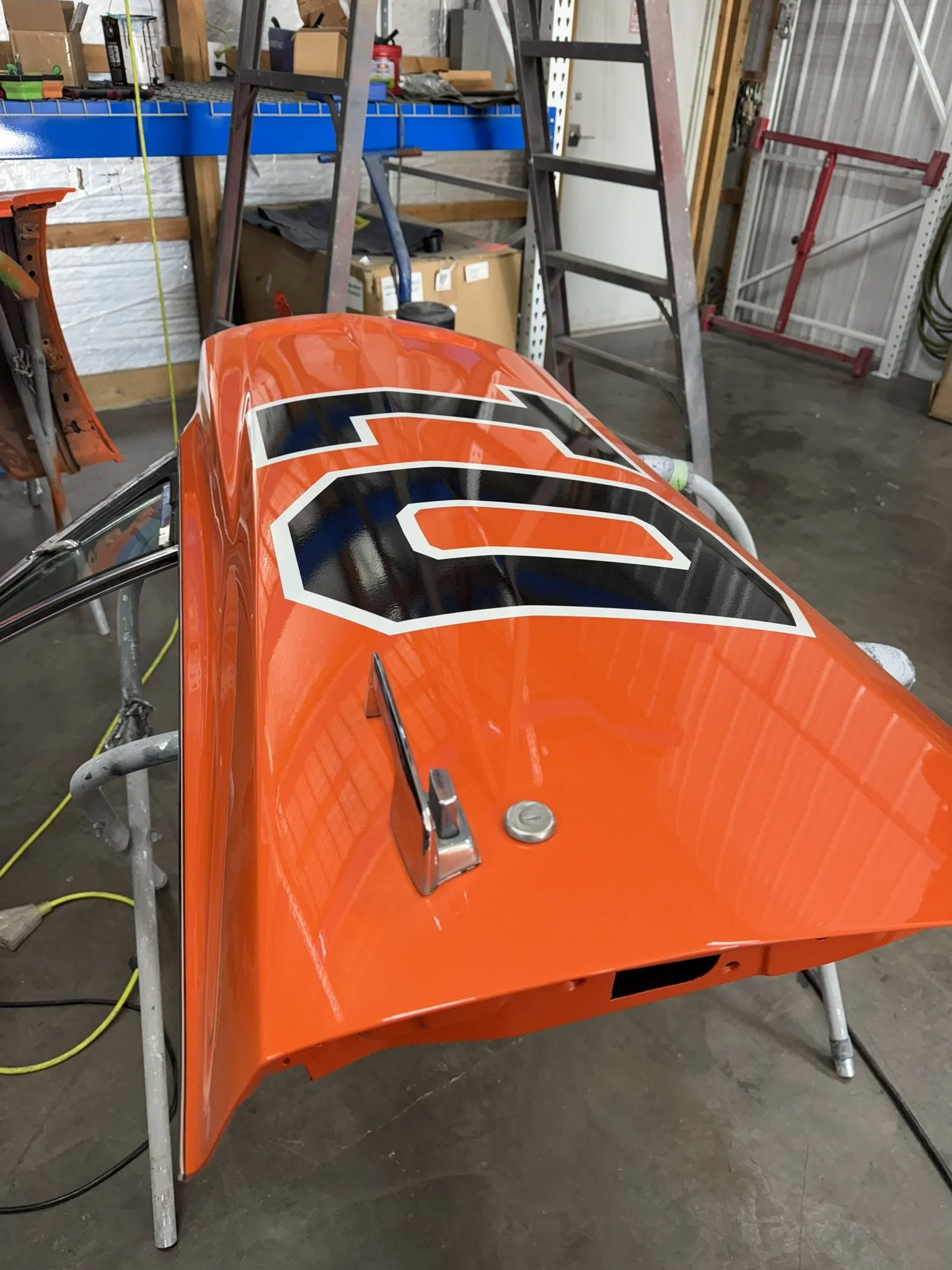 A bright orange race car body with black and white graphics, mounted on a metal stand in a garage workspace.