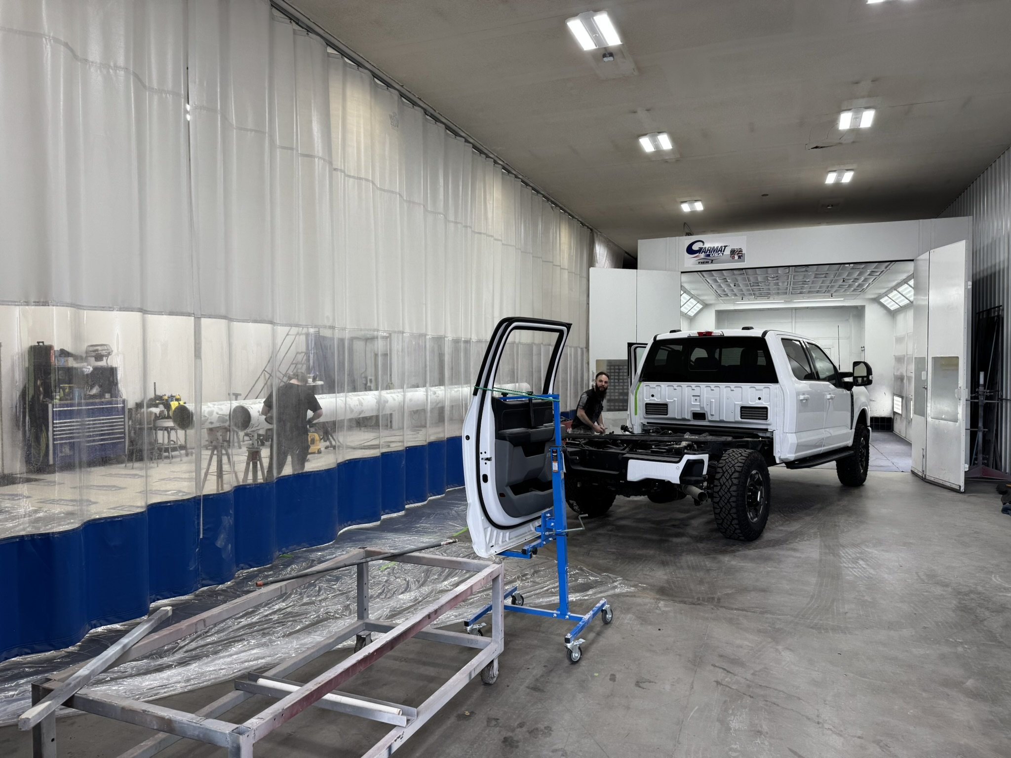 An indoor vehicle repair or manufacturing shop with a white pickup truck missing its front panels, a person working on it, a blue tool cart, and a clear plastic curtain wall on the left side.