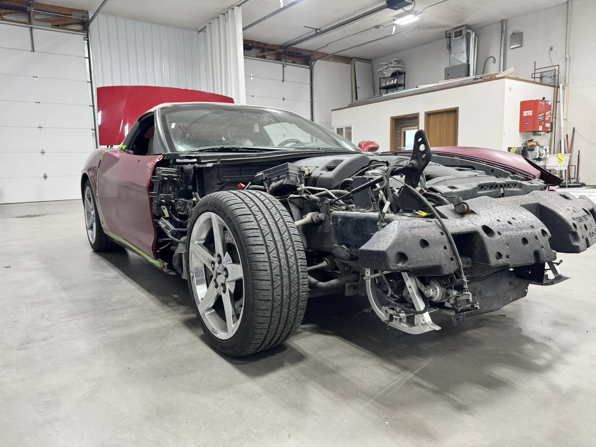 A red sports car inside a garage with the hood open and the front bumper removed, exposing the engine and internal parts of the vehicle, with a storage room and tools in the background.