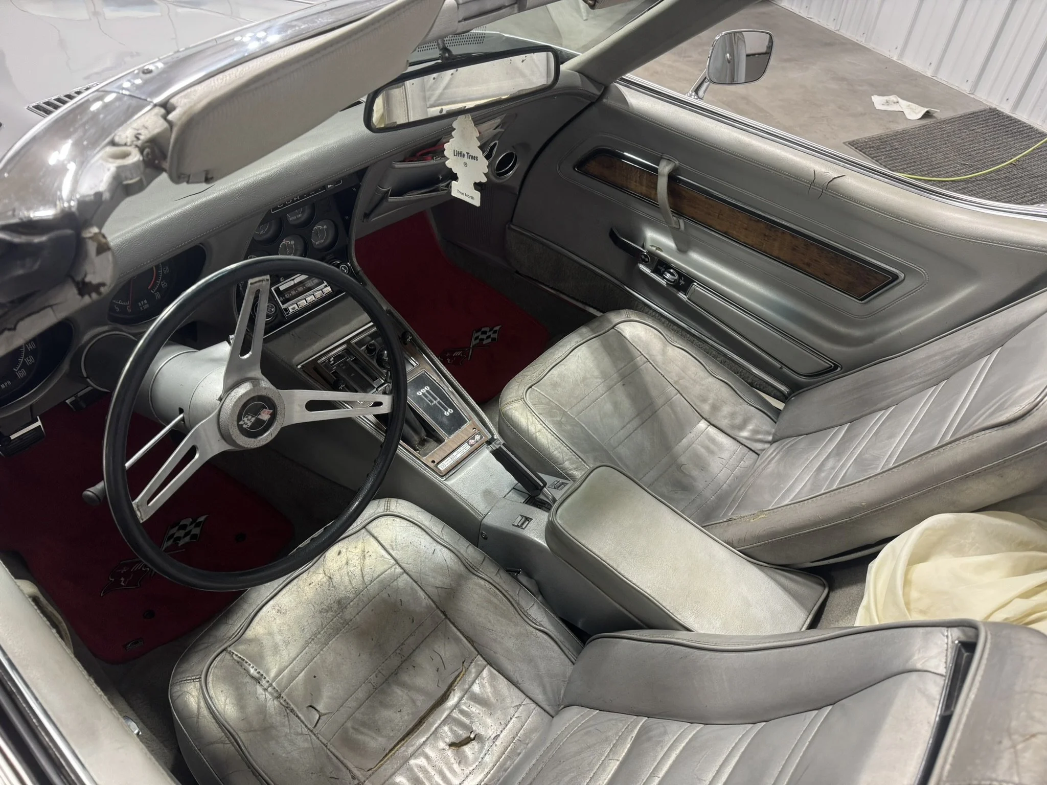 The interior of a weathered vintage car with worn gray leather seats, a wooden-trimmed dashboard, a steering wheel with a Chevrolet emblem, and an air freshener hanging from the rearview mirror.