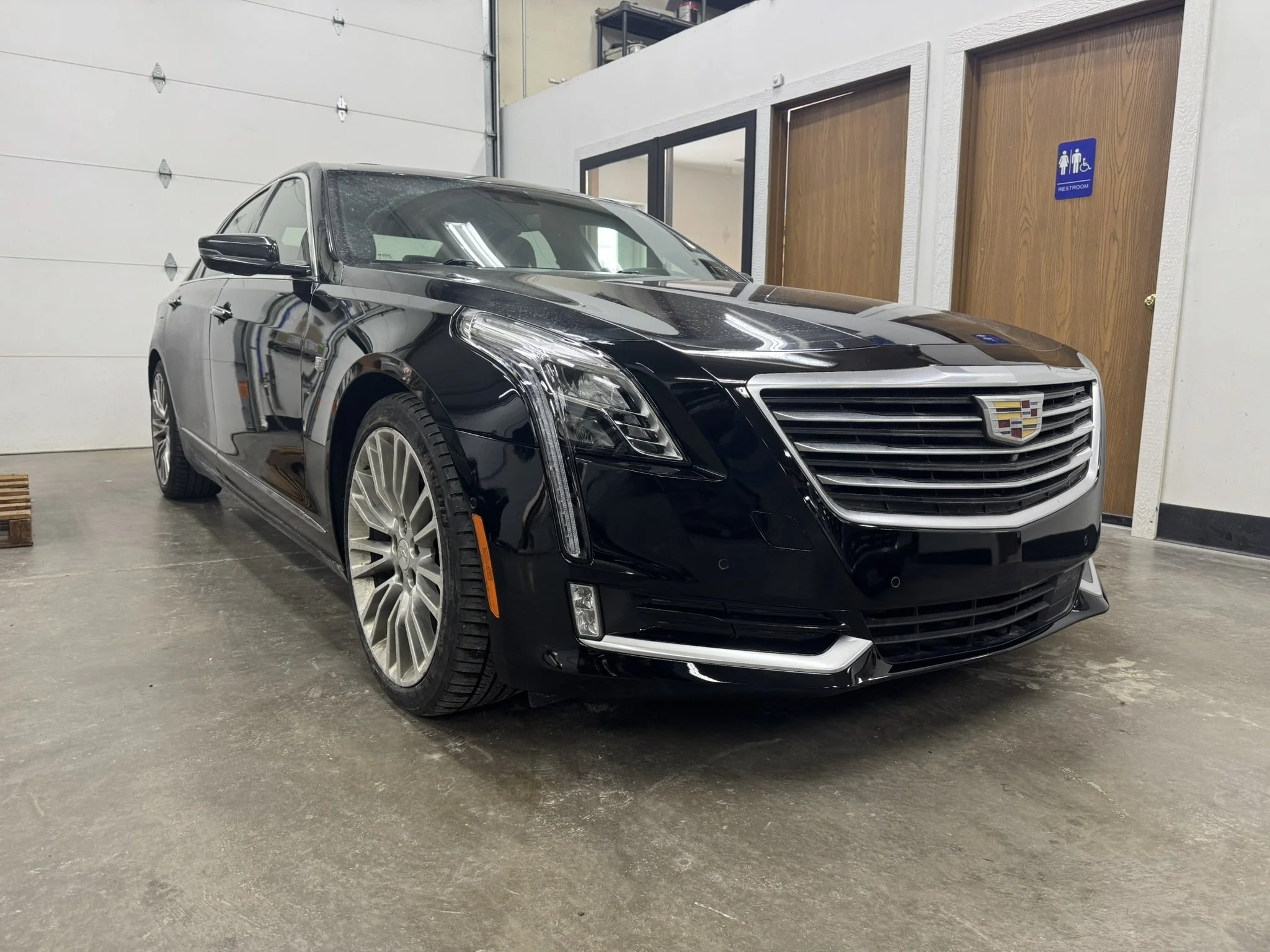 A black Cadillac sedan parked inside a garage with a concrete floor. The garage has white painted walls, a large white roll-up garage door, and two wooden doors with a restroom sign."