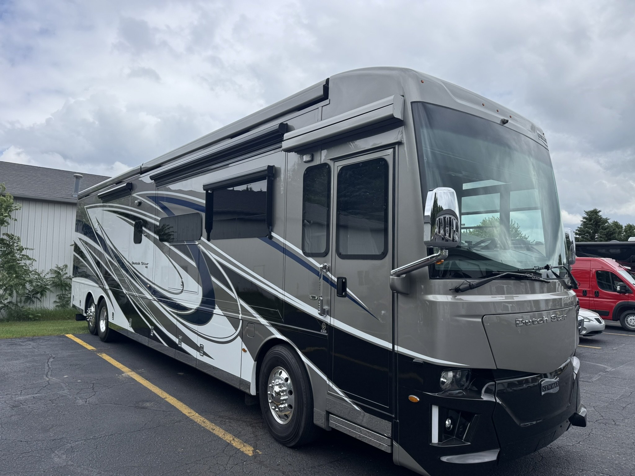 A modern motorhome parked in a lot with a building and other vehicles in the background. The motorhome has a sleek design with black, gray, and white graphics on its side.