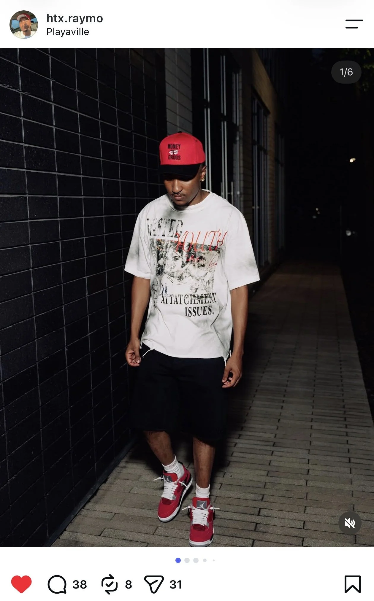A young man with dark skin standing on a wooden pathway at night, wearing a red and black baseball cap, a graphic white T-shirt, black shorts, and red and white sneakers.