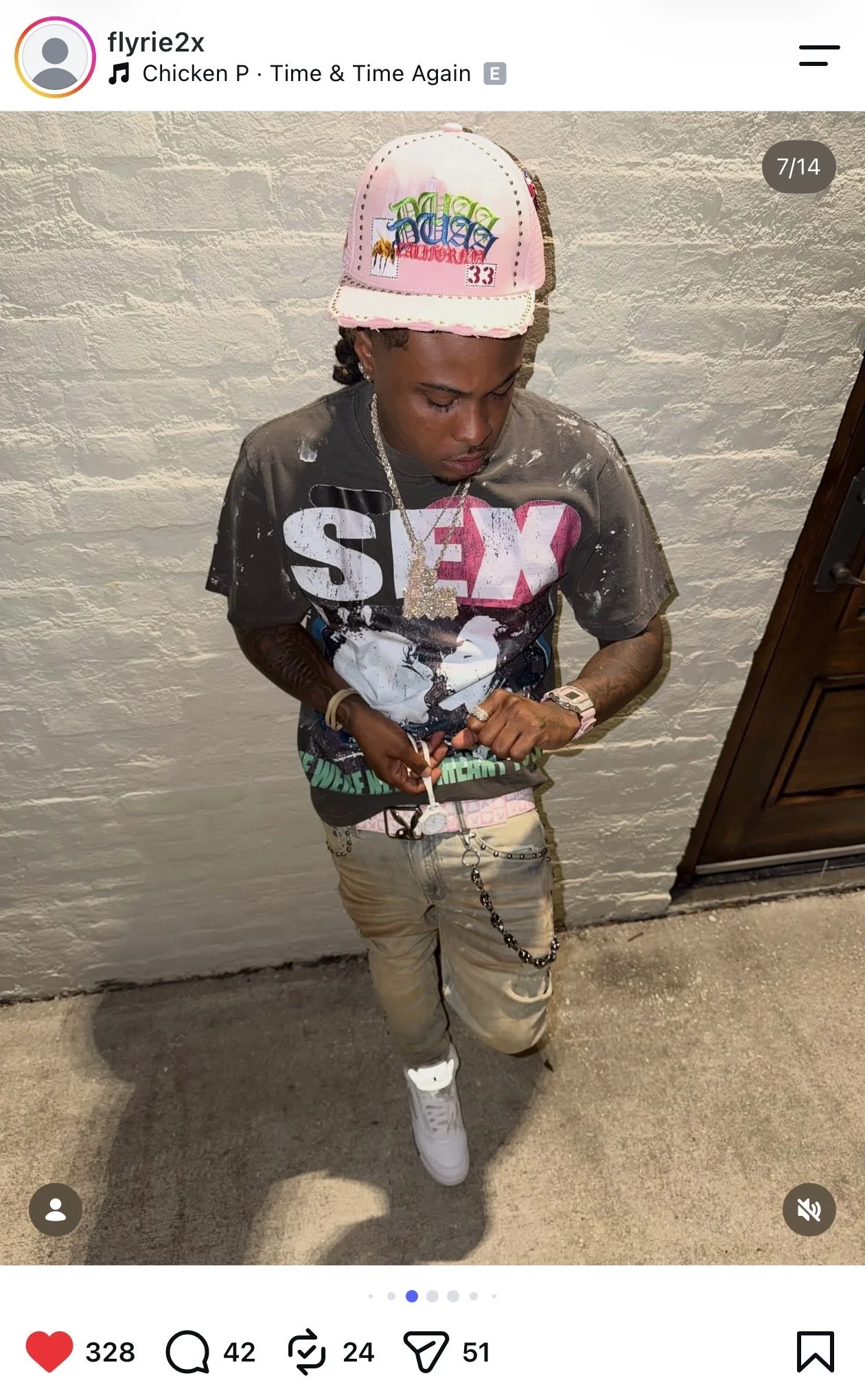 A young man wearing a pink and white baseball cap, layered chains, a black graphic T-shirt with a large face and red and white text, beige distressed pants, and white sneakers, standing against a textured white wall and looking down, with his hands a