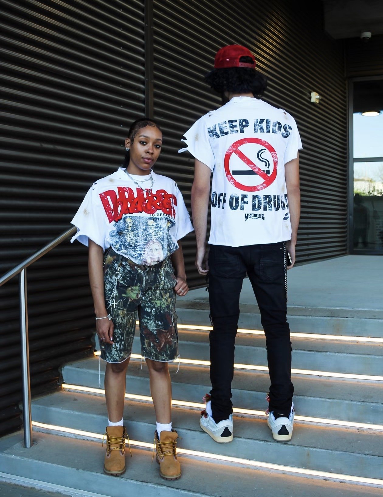 Two young people, a woman and a man, standing on steps outside. The woman faces forward, wearing a graphic T-shirt with edgy text and camo shorts, and tan boots. The man faces away, wearing a white T-shirt with a no-smoking sign and the words 'Keep Kids Off of Drugs,' black pants, white sneakers, and a red cap.