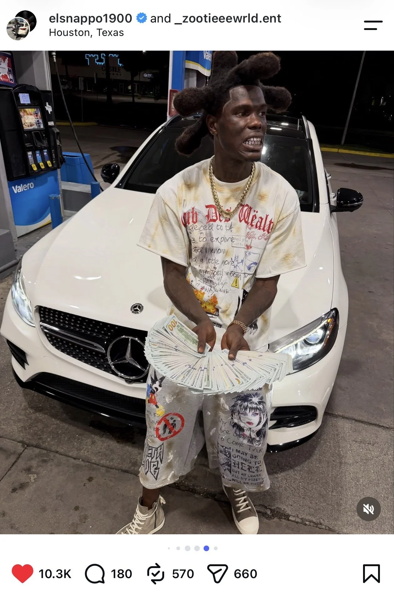 Man with styled dreadlocks holding a fan of cash stands in front of a white Mercedes-Benz car at a gas station during nighttime in Houston, Texas.