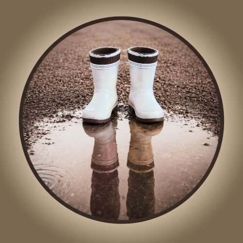 White rain boots with black and white striped tops reflected in a puddle on a gravel surface.