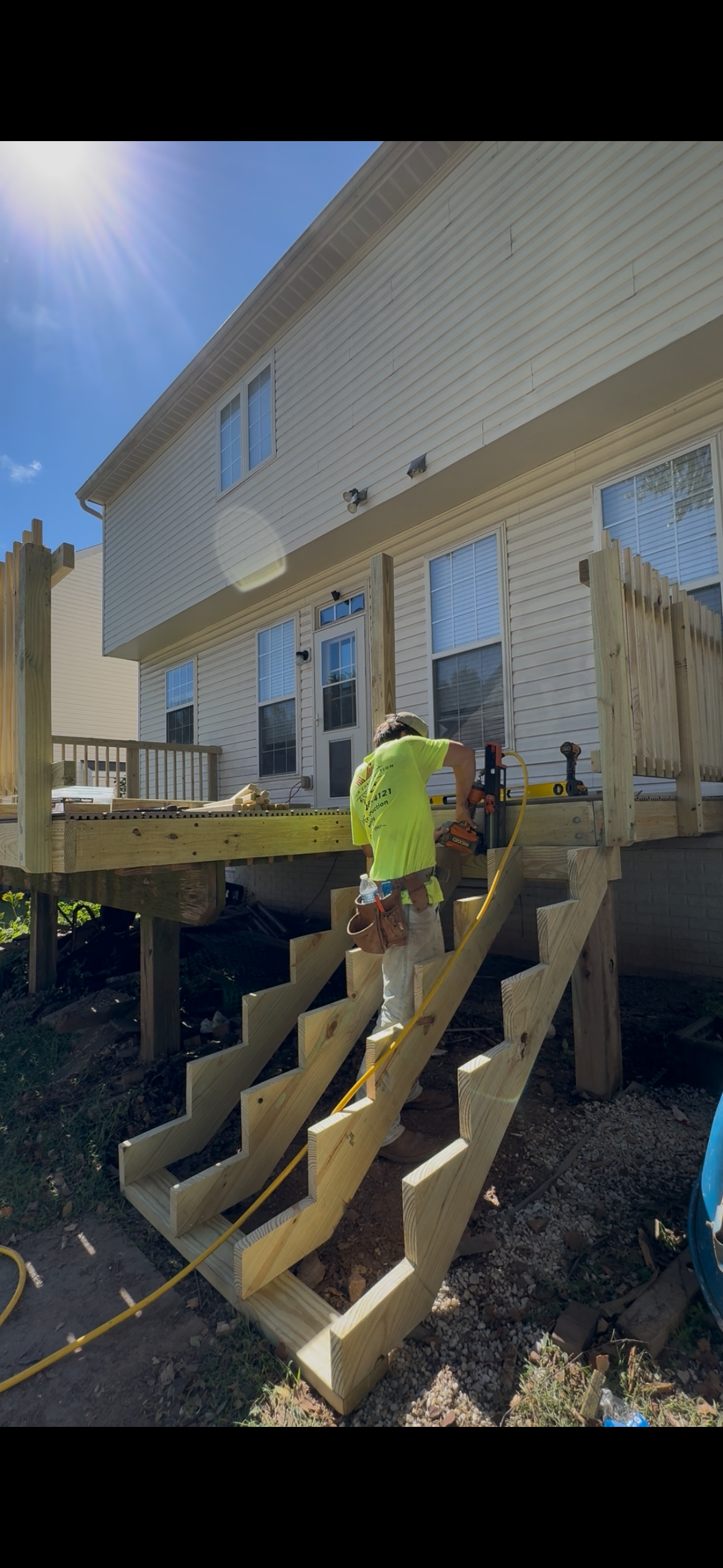 A construction worker in a bright yellow shirt is installing a wooden staircase outside a beige house with vinyl siding. The worker is using tools on the stairs, which are in the process of being built, with sunlight shining from the top left corner.