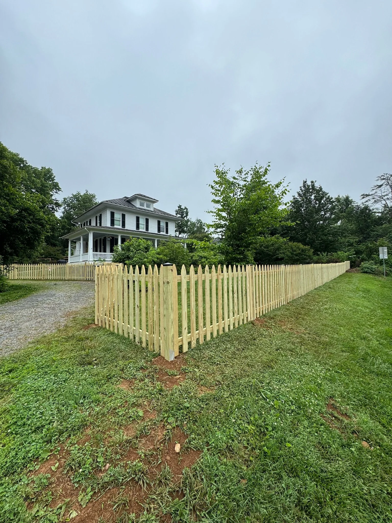 A white two-story house with black shutters, surrounded by a beige picket fence and green trees, under a cloudy sky.