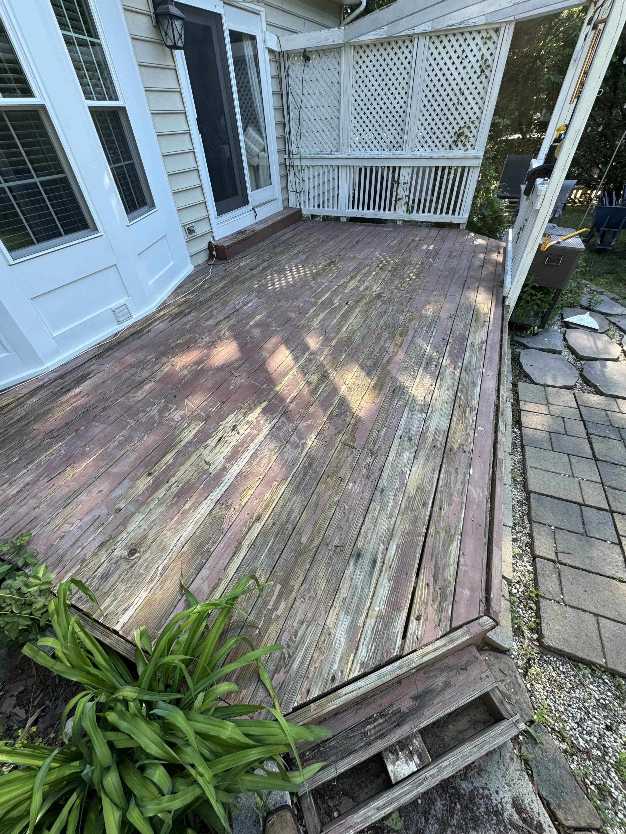 A wooden deck outside a house with a lattice privacy fence, a sliding glass door, potted plants, and a garden pathway with paving stones.