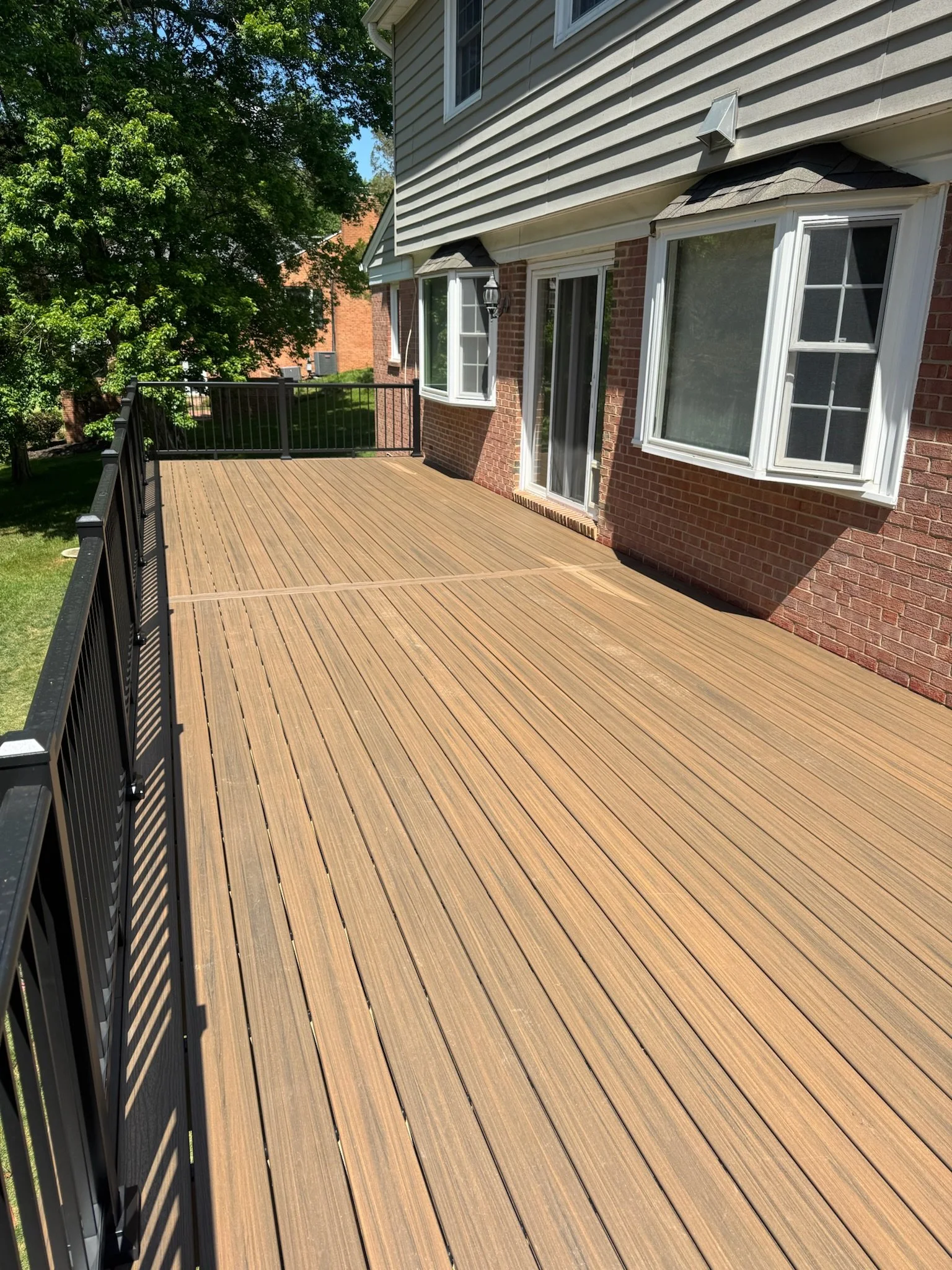 View of a wooden deck attached to a house with white siding and brick exterior, surrounded by green trees and grass.